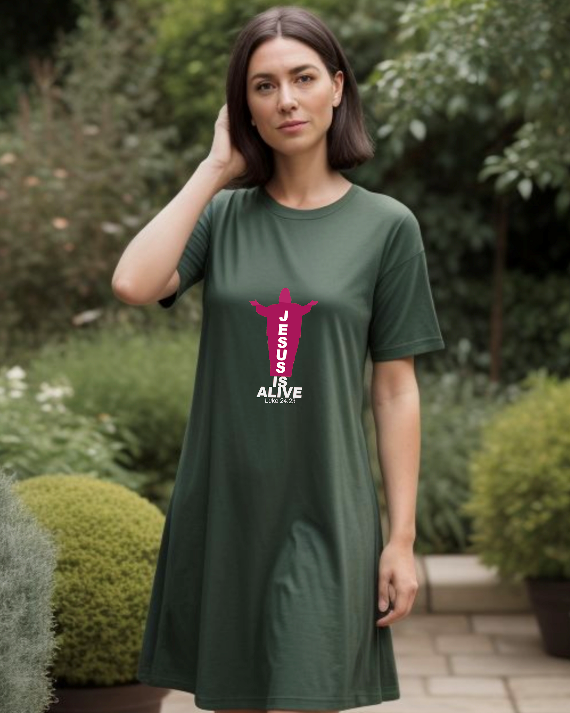 A woman in a garden wearing a dark green Heavenly Dreamwear t-shirt dress with a red and white logo of a silhouette of Jesus with the name "JESUS" embedded in it. Underneath is the text "IS ALIVE" with the scriptural reference: Luke 24:23.