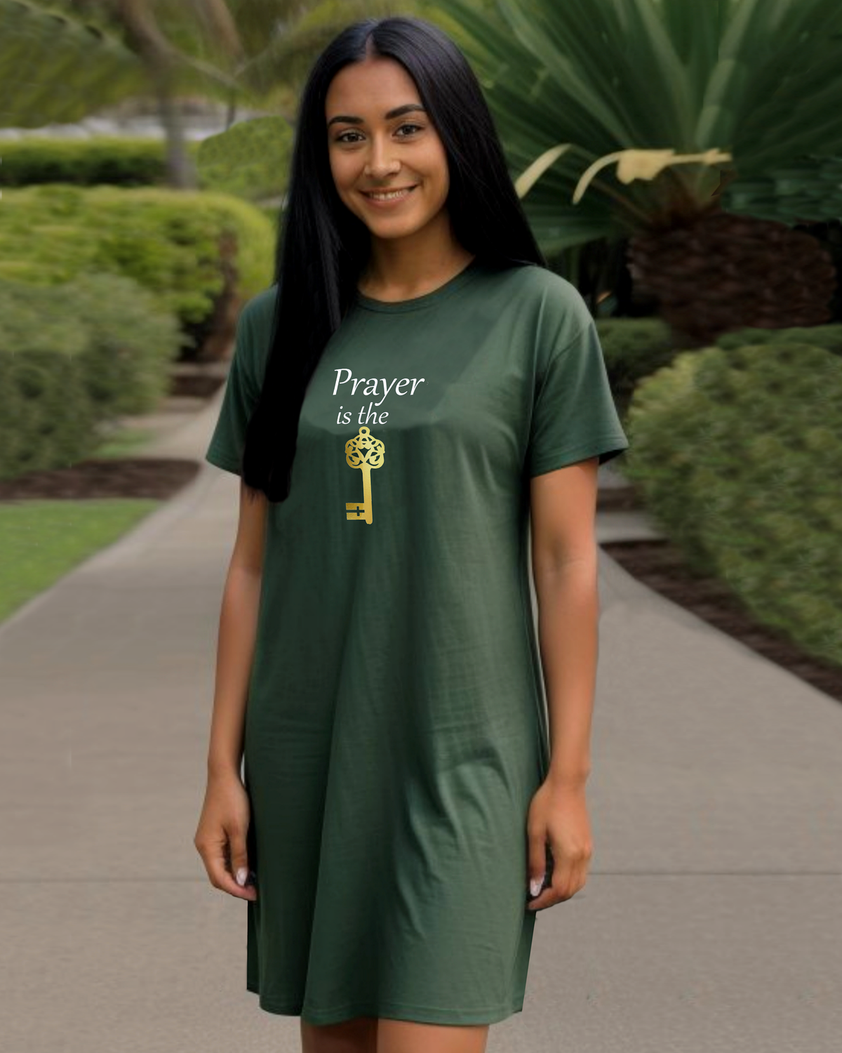 A woman wearing a dark green t-shirt dress with a graphic that reads 'Prayer is the key' in white and gold, standing on a paved path in a landscaped garden.