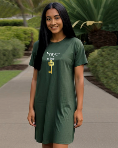 A woman wearing a dark green t-shirt dress with a graphic that reads 'Prayer is the key' in white and gold, standing on a paved path in a landscaped garden.