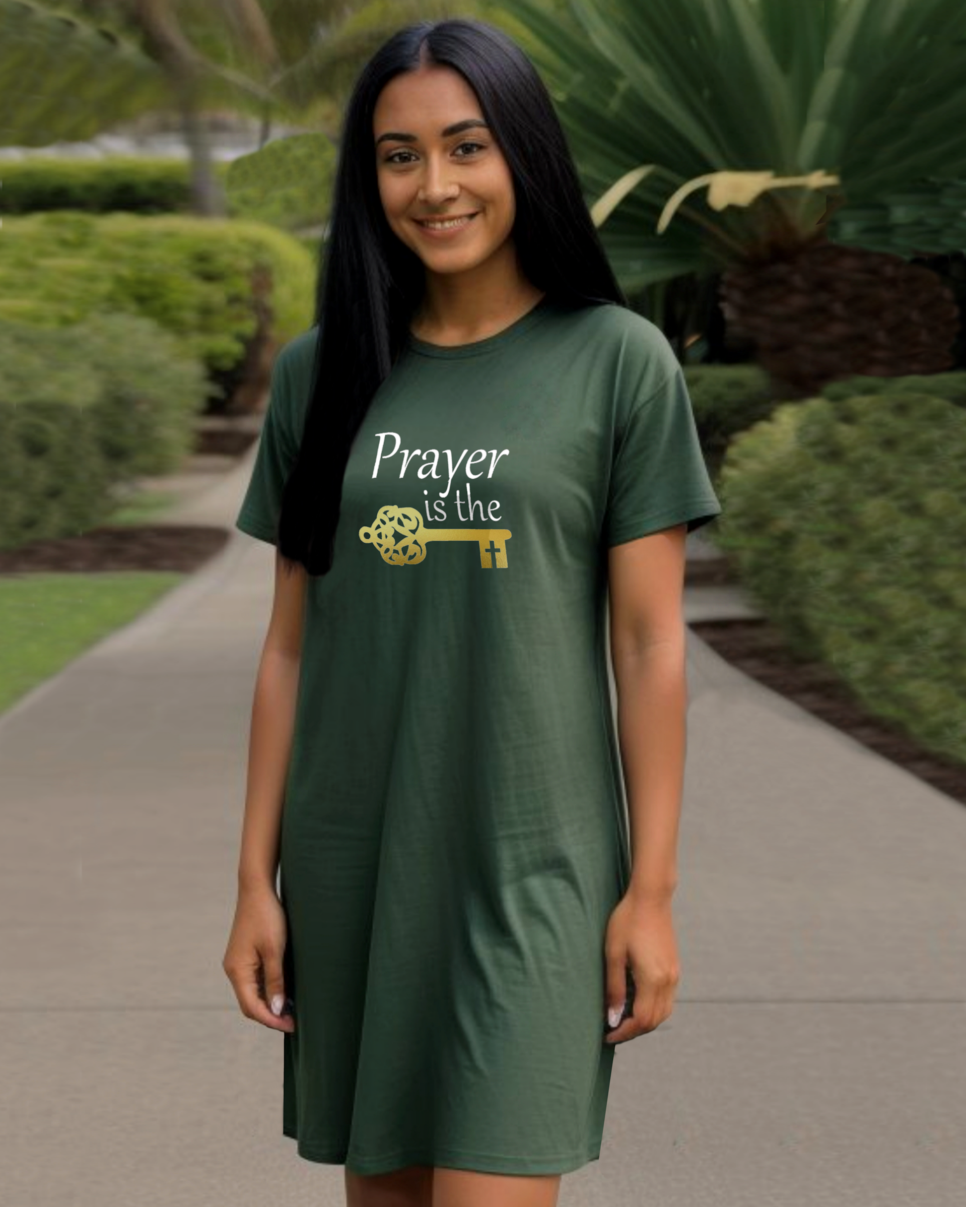 A woman in a garden wearing a dark green t-shirt dress featuring a logo design by Heavenly Dreamwear.  The logo includes white text that reads, "Prayer is the," followed by a gold key symbol.