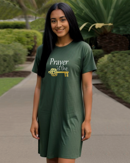 A woman in a garden wearing a dark green t-shirt dress featuring a logo design by Heavenly Dreamwear.  The logo includes white text that reads, "Prayer is the," followed by a gold key symbol.