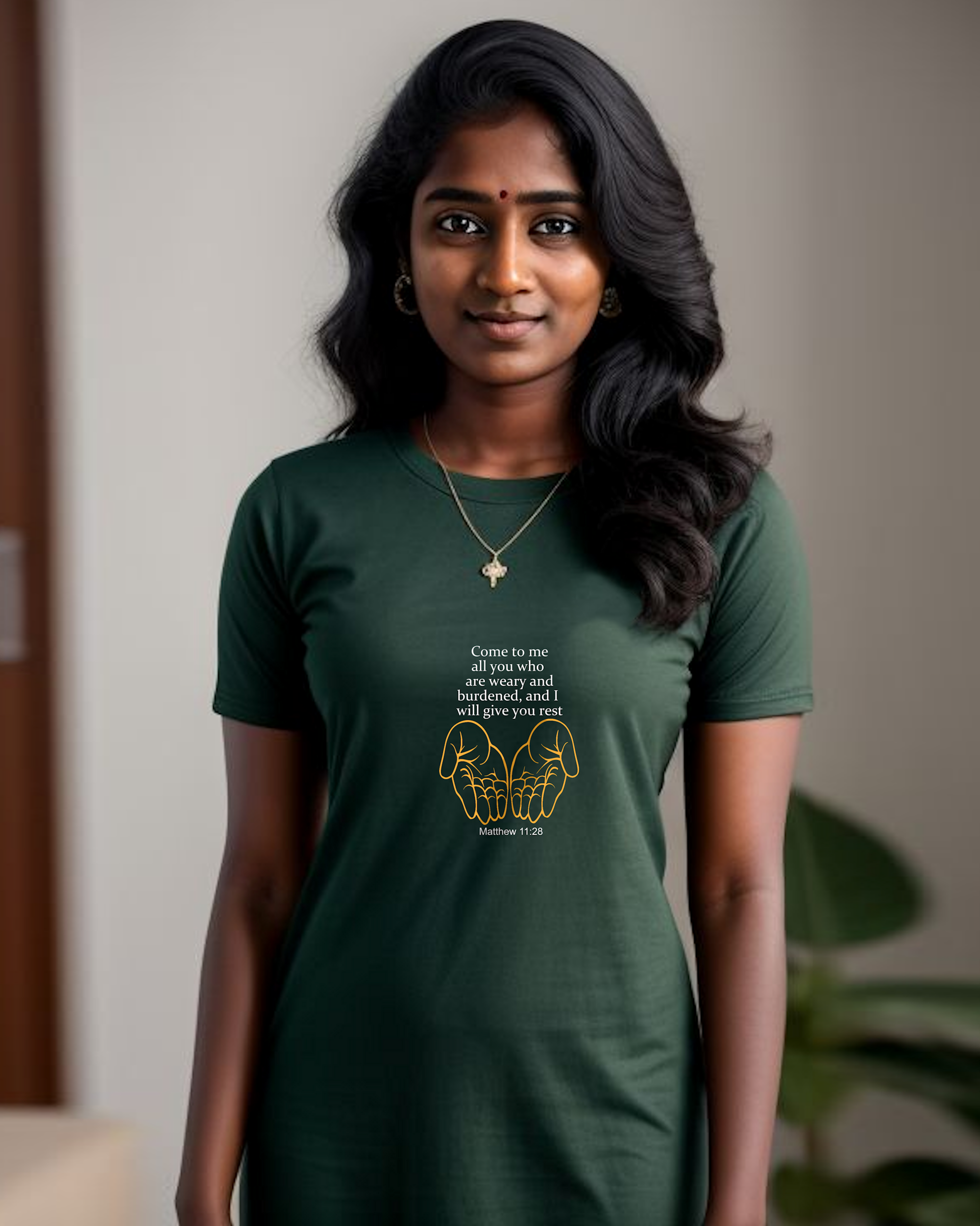 A woman standing indoors wearing a dark green Heavenly Dreamwear t-shirt dress with white "come to me all you who are weary" text with two yellow hands facing palm up logo printed on the front.