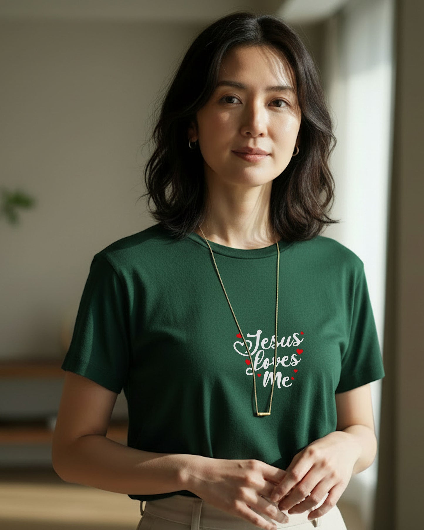 A woman indoors wearing a dark green t-shirt with white 'Jesus Loves Me' text with red hearts and beige pants