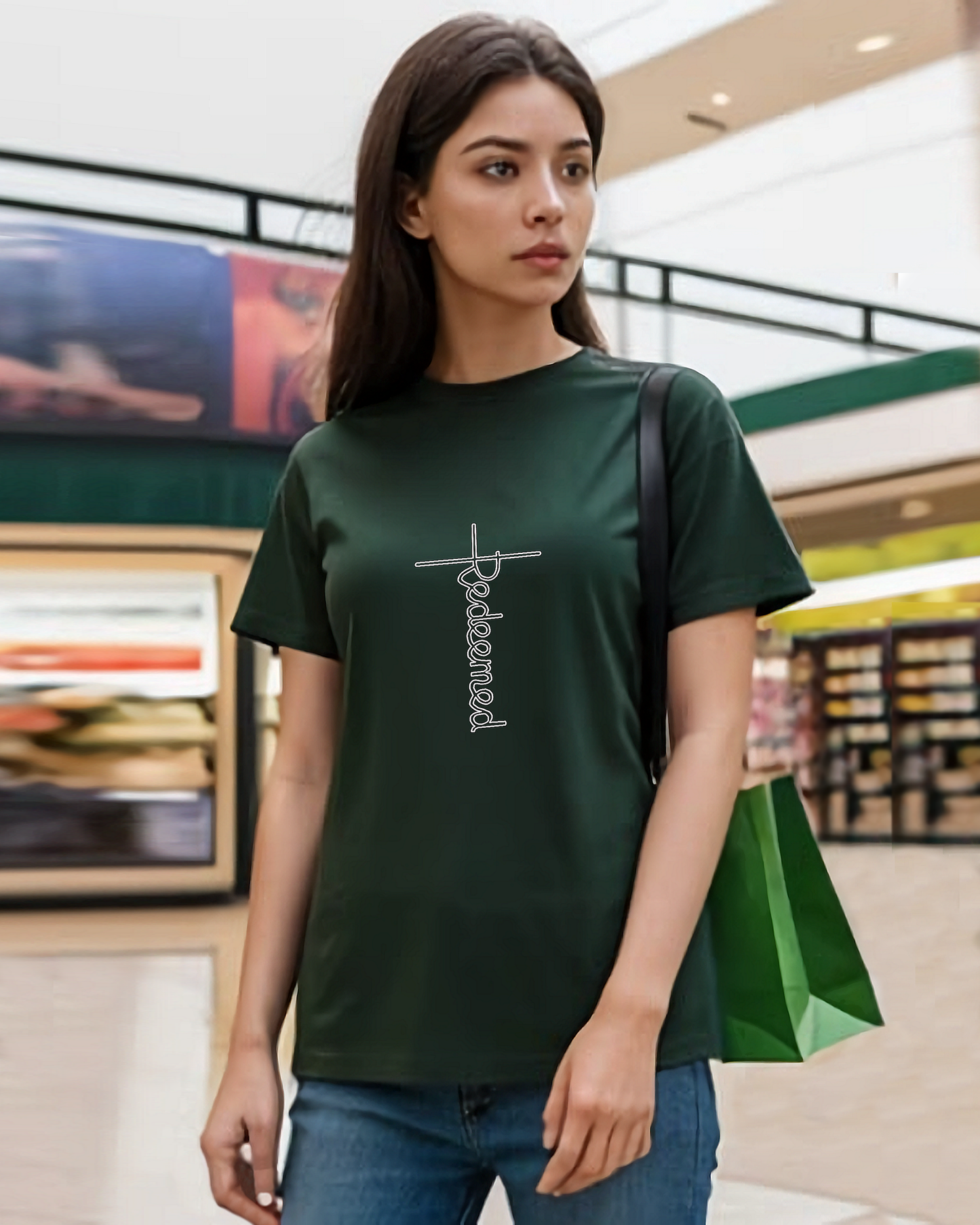A woman standing in a shopping centre wearing a dark green t-shirt with an elaborate  Heavenly Dreamwear designed 'Redeemed' logo printed on the front.