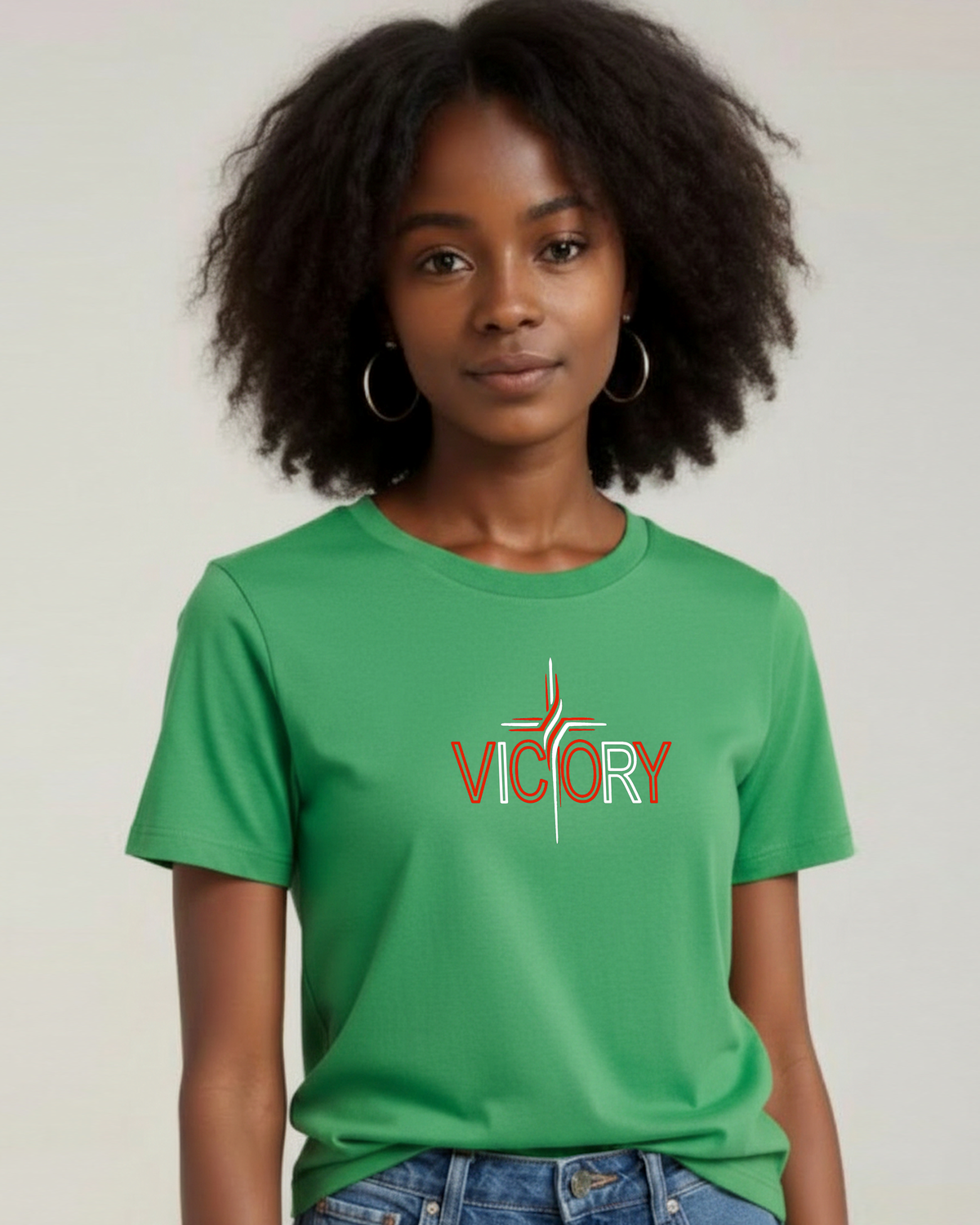 A woman with dark curly hair wearing a green short-sleeve t-shirt with a 'VICTORY' graphic on the chest, paired with blue jeans, against a plain light background.
