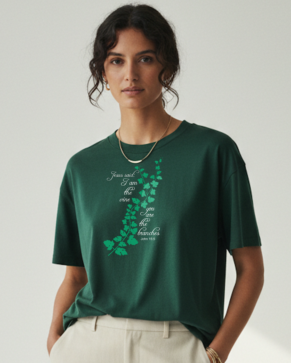 A woman wearing a dark green short-sleeve t-shirt with a light green vine graphic and white script text 'Jesus said I am the vine you are the branches' on the front, paired with off-white trousers, against a plain light gray background.