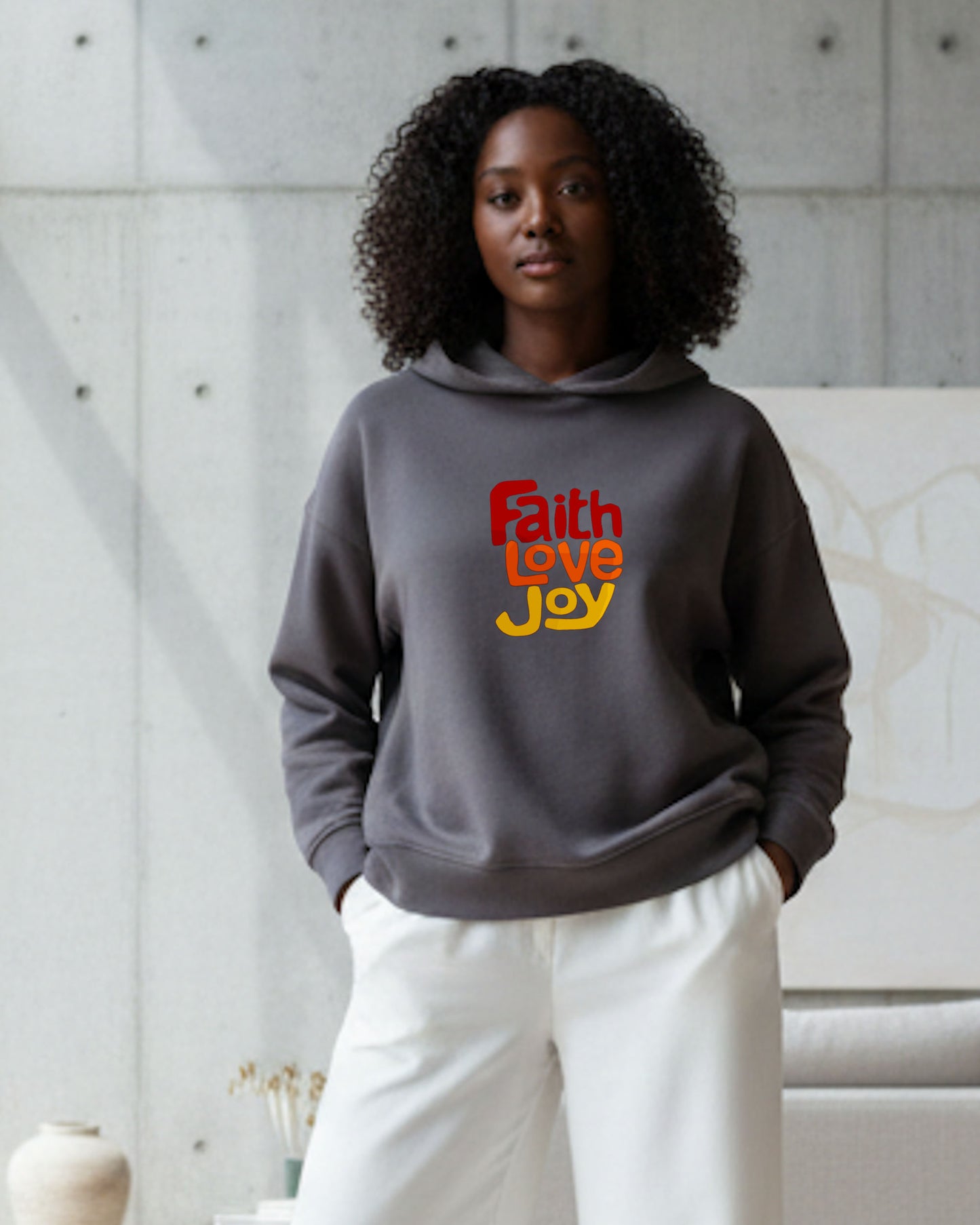 Woman wearing a gray hoodie with 'Faith Love Joy' text in red, orange and yellow, standing against a neutral background.