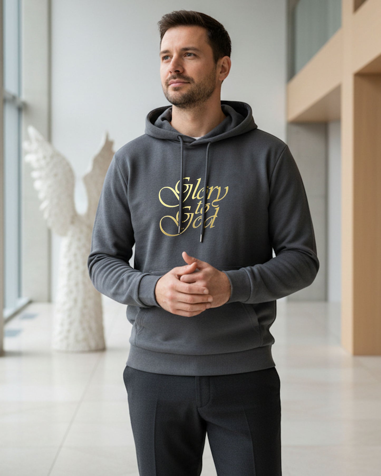 Man standing indoors wearing a dark grey hoodie with a gold Heavenly Dreamwear "Glory to God" text logo printed on front. 
