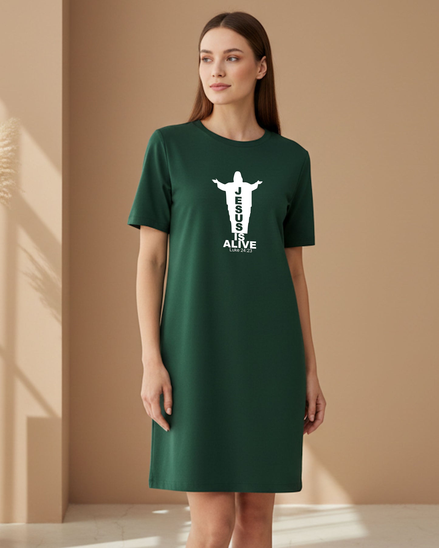 A woman indoors wearing a dark green Heavenly Dreamwear t-shirt dress with a white logo of a silhouette of Jesus with the name "JESUS" embedded in it. Underneath is the text "IS ALIVE" with the scriptural reference: Luke 24:23.