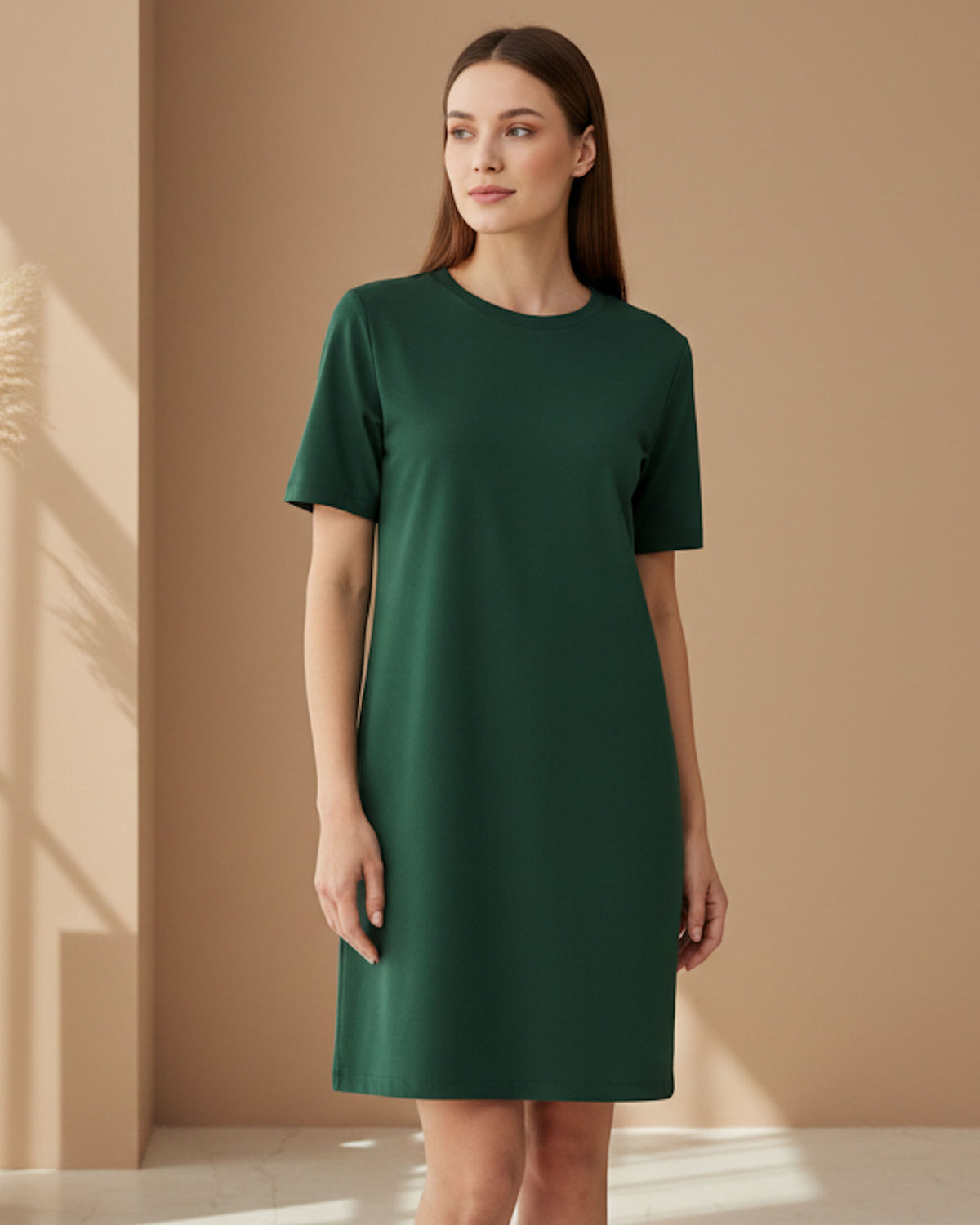 A woman modeling a short-sleeve, knee-length dark green dress in a minimalist studio setting with warm beige walls and soft natural shadows.