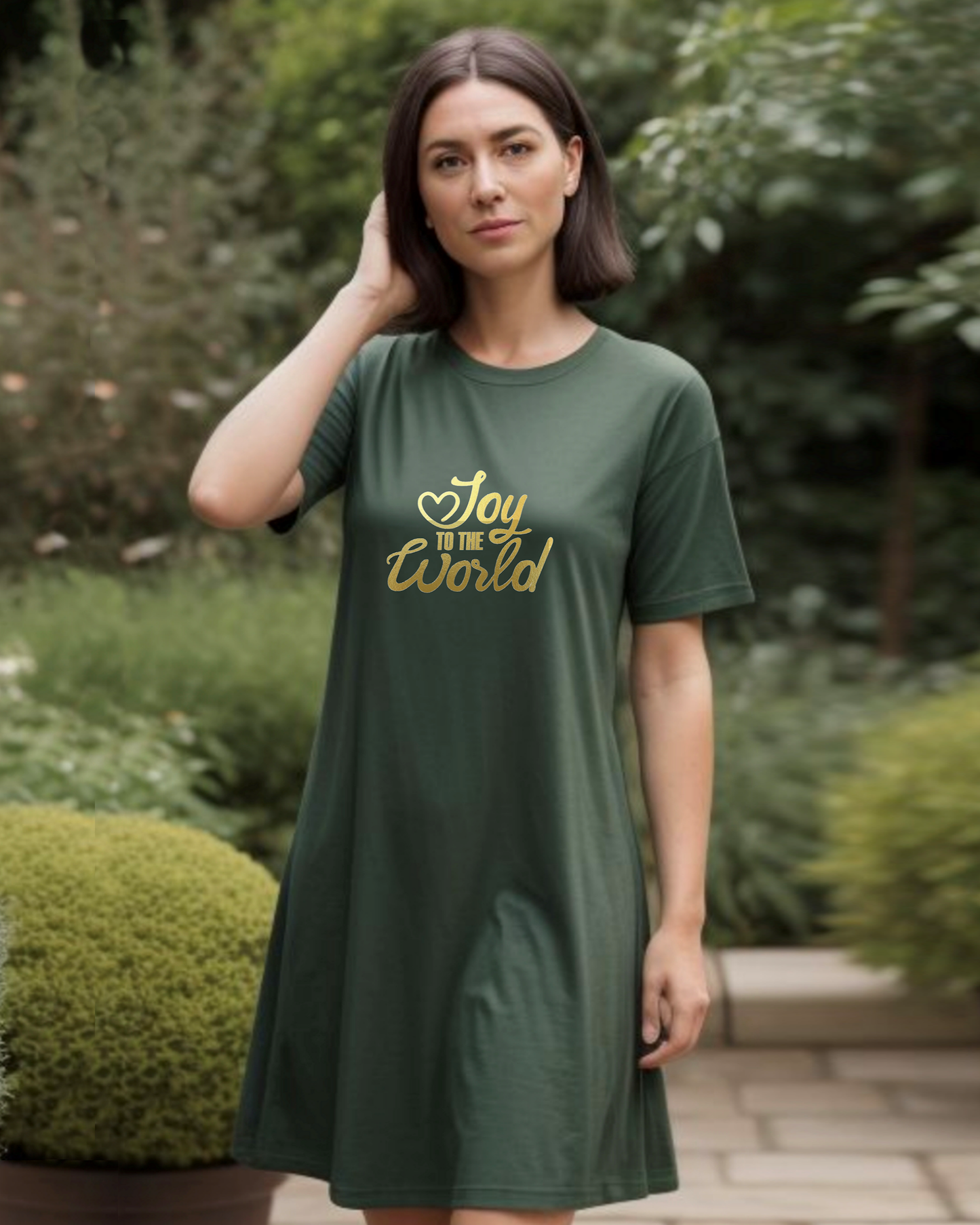 A woman standing in a garden wearing a dark green Heavenly Dreamwear t-shirt dress with a gold "Joy to the World" logo printed on the front.