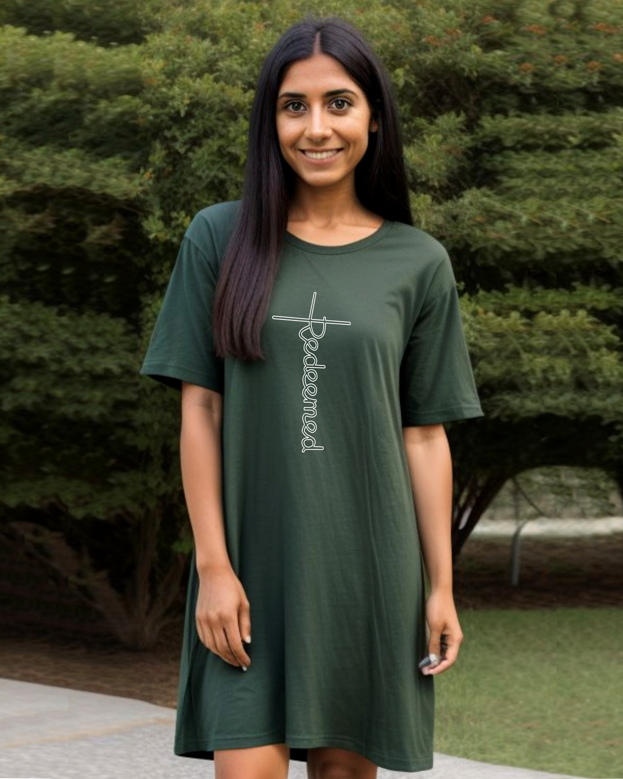 A woman in a garden wearing a green Heavenly Dreamwear t-shirt dress with an elaborate white 'Redeemed' logo printed on the front. 