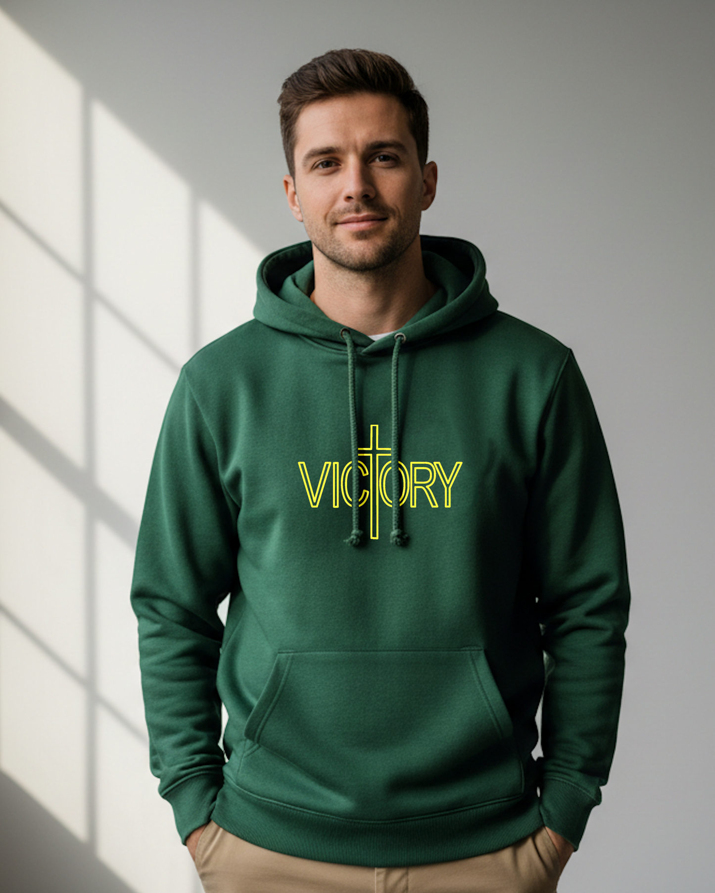 A man wearing a dark green pullover hoodie with a yellow 'VICTORY' graphic and cross on the chest, standing against a gray wall with window shadow light.