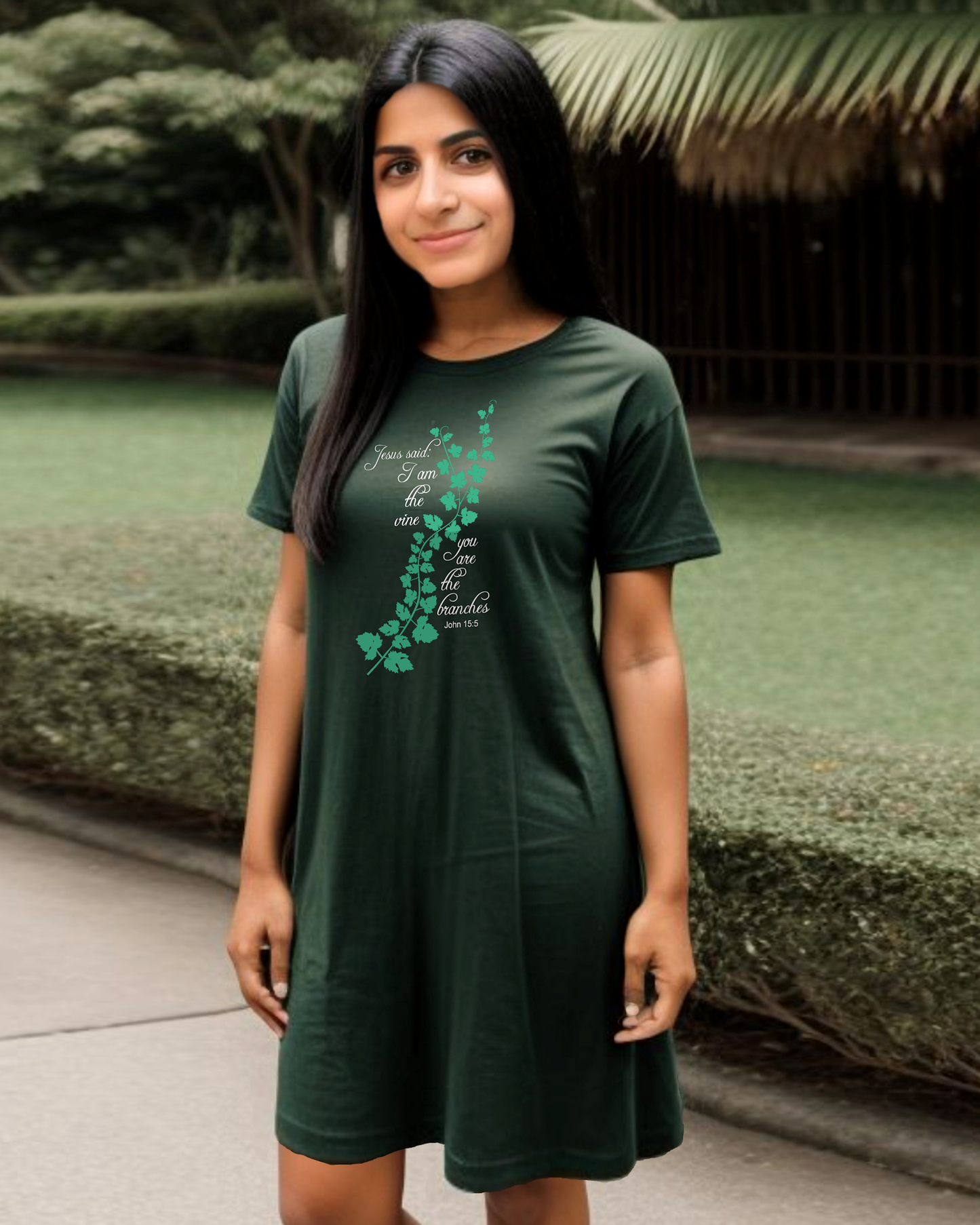 Woman standing in a garden wearing a dark green t-shirt dress with a logo made up of a green vine draped across the front with a biblical scripture on either side in white text from John 15:5