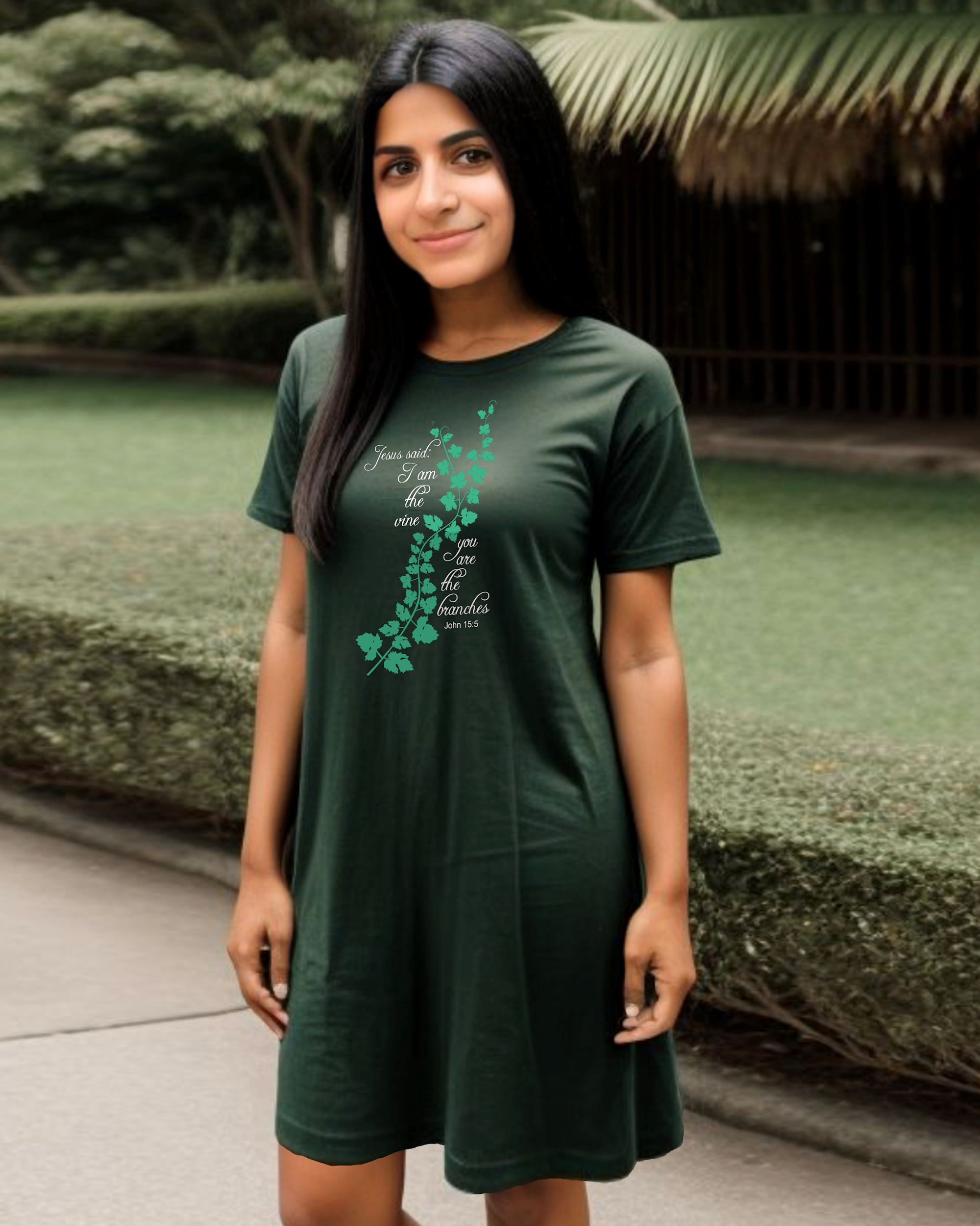 Woman standing in a garden wearing a dark green t-shirt dress with a logo made up of a green vine draped across the front with a biblical scripture on either side in white text from John 15:5