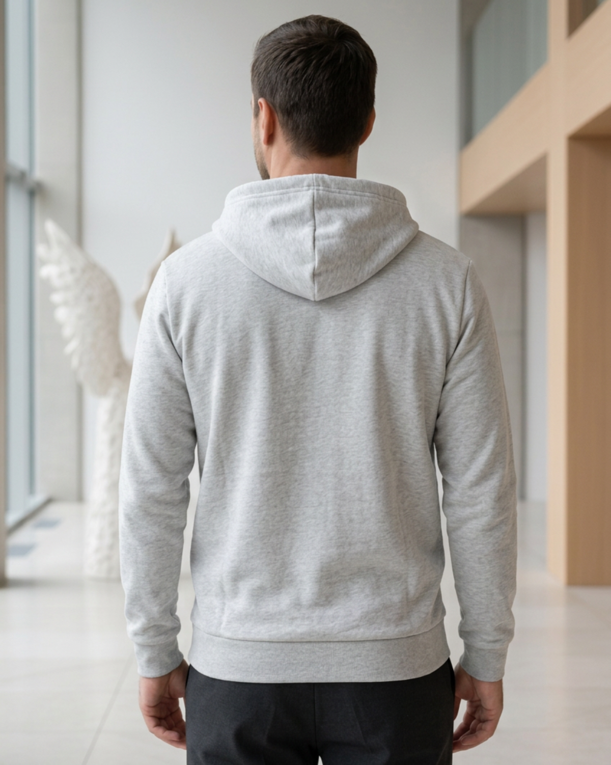 A man viewed from behind wearing a dark gray pullover hoodie and dark pants, standing in a modern indoor hallway with light-colored walls and architectural columns.