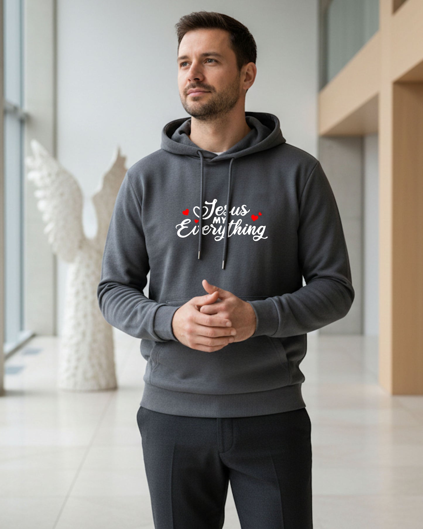 Man wearing a gray hoodie with a Christian faith inspired text logo saying "Jesus My Everyhing" with red hearts designed by Heavenly Dreamwear printed on the front in a modern indoor setting