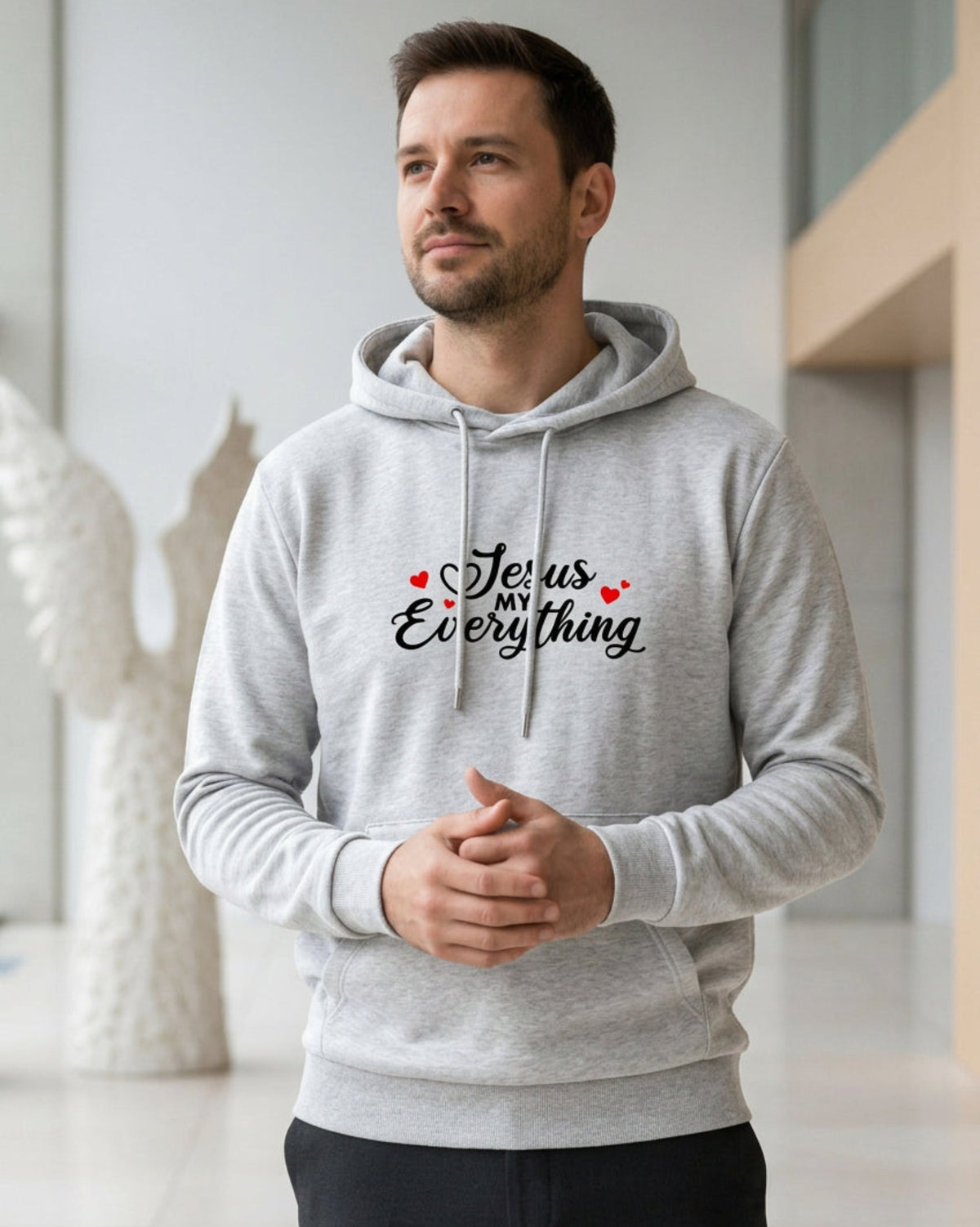 Man wearing a gray hoodie with a Christian faith inspired text logo saying "Jesus My Everyhing" with red hearts designed by Heavenly Dreamwear printed on the front in a modern indoor setting