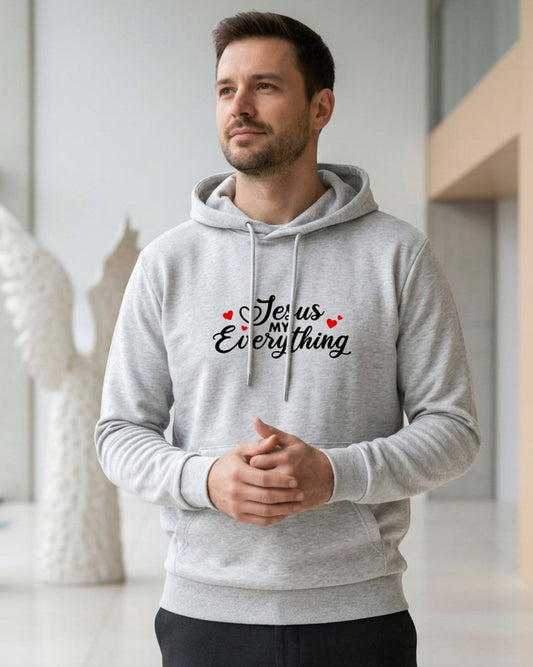 Man wearing a gray hoodie with a Christian faith inspired text logo saying "Jesus My Everyhing" with red hearts designed by Heavenly Dreamwear printed on the front in a modern indoor setting