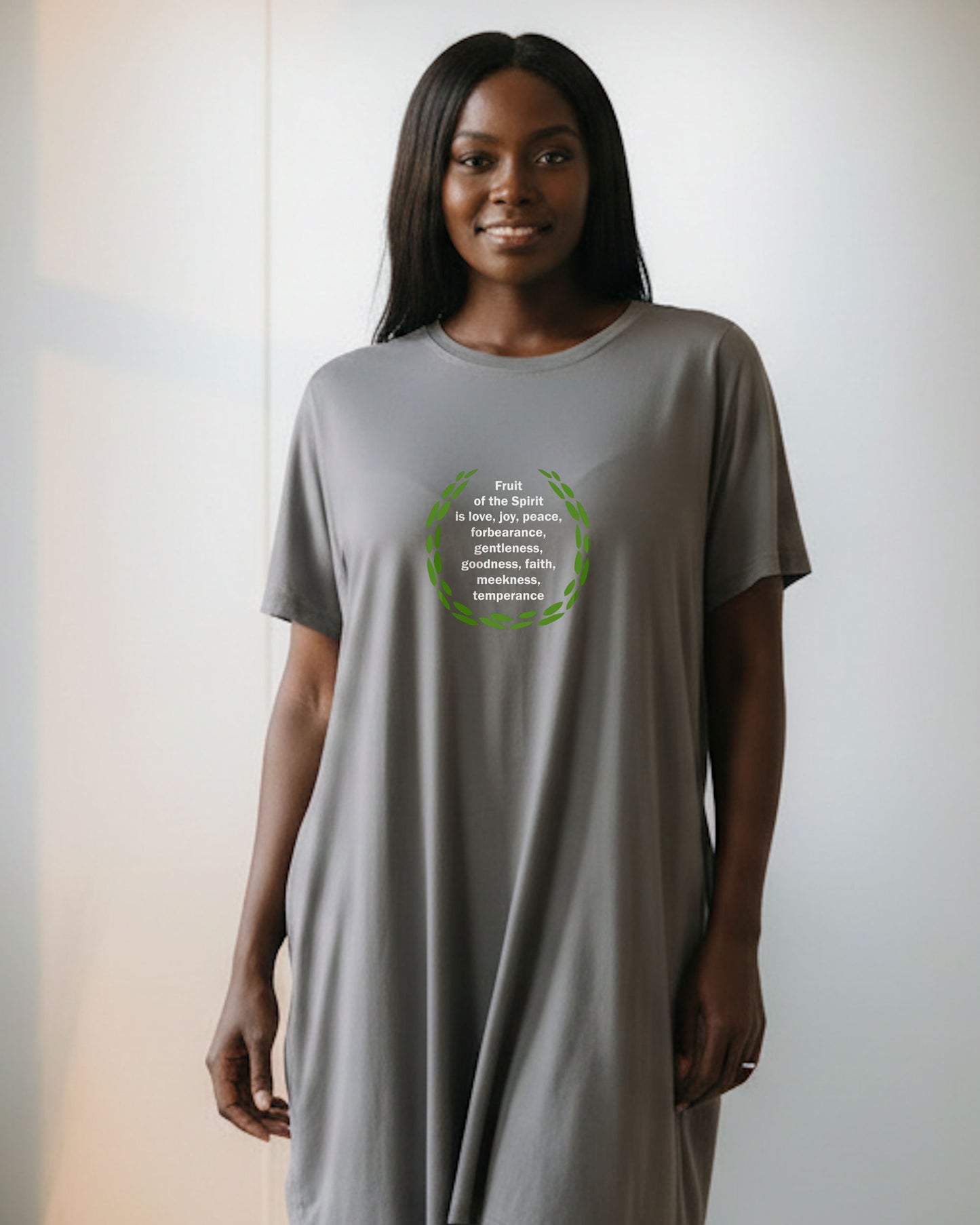 A woman indoors wearing a grey t-shirt dress with a logo stating the fruit of the Holy Spirit in white text surrounded by green leaves.