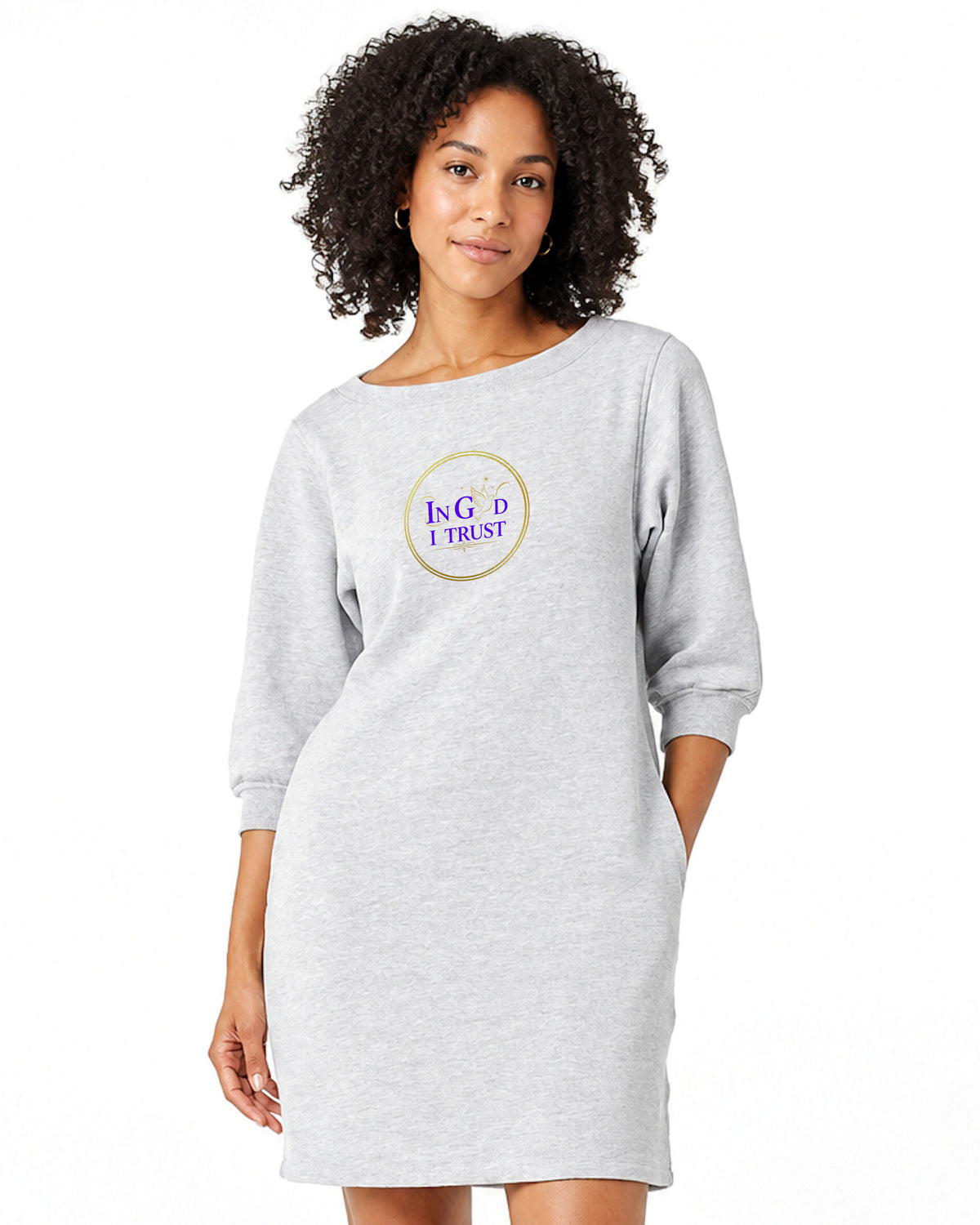 A woman wearing a light gray heather boat neck sweatshirt dress with a circular logo in gold and purple that reads, "In God I Trust" against a white bakground.