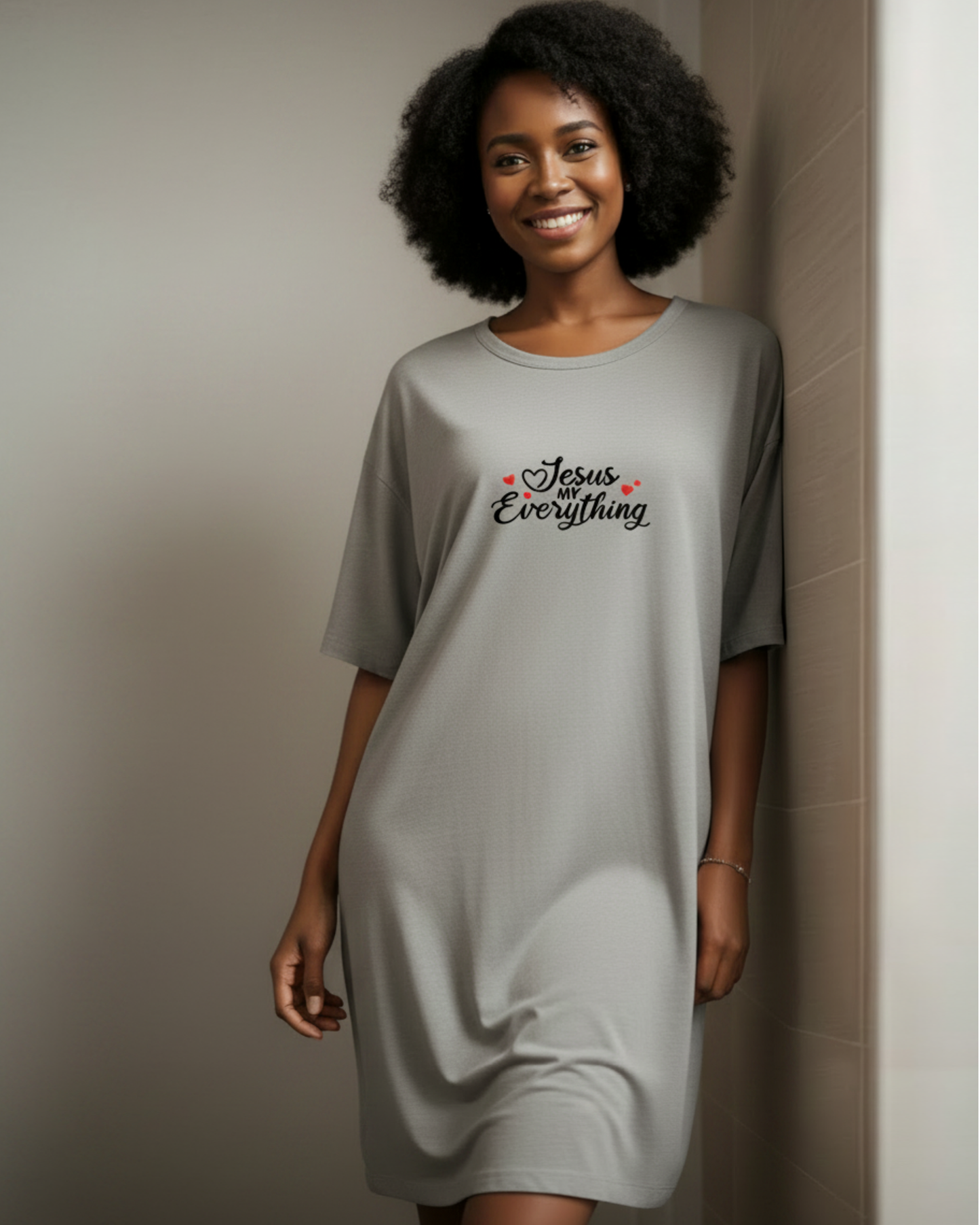 A woman wearing a light gray oversized t-shirt dress with 'Jesus Everything' printed in black script with small red hearts on the chest, standing against a neutral beige tiled wall.