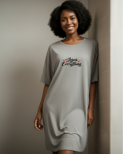 A woman wearing a light gray oversized t-shirt dress with 'Jesus Everything' printed in black script with small red hearts on the chest, standing against a neutral beige tiled wall.