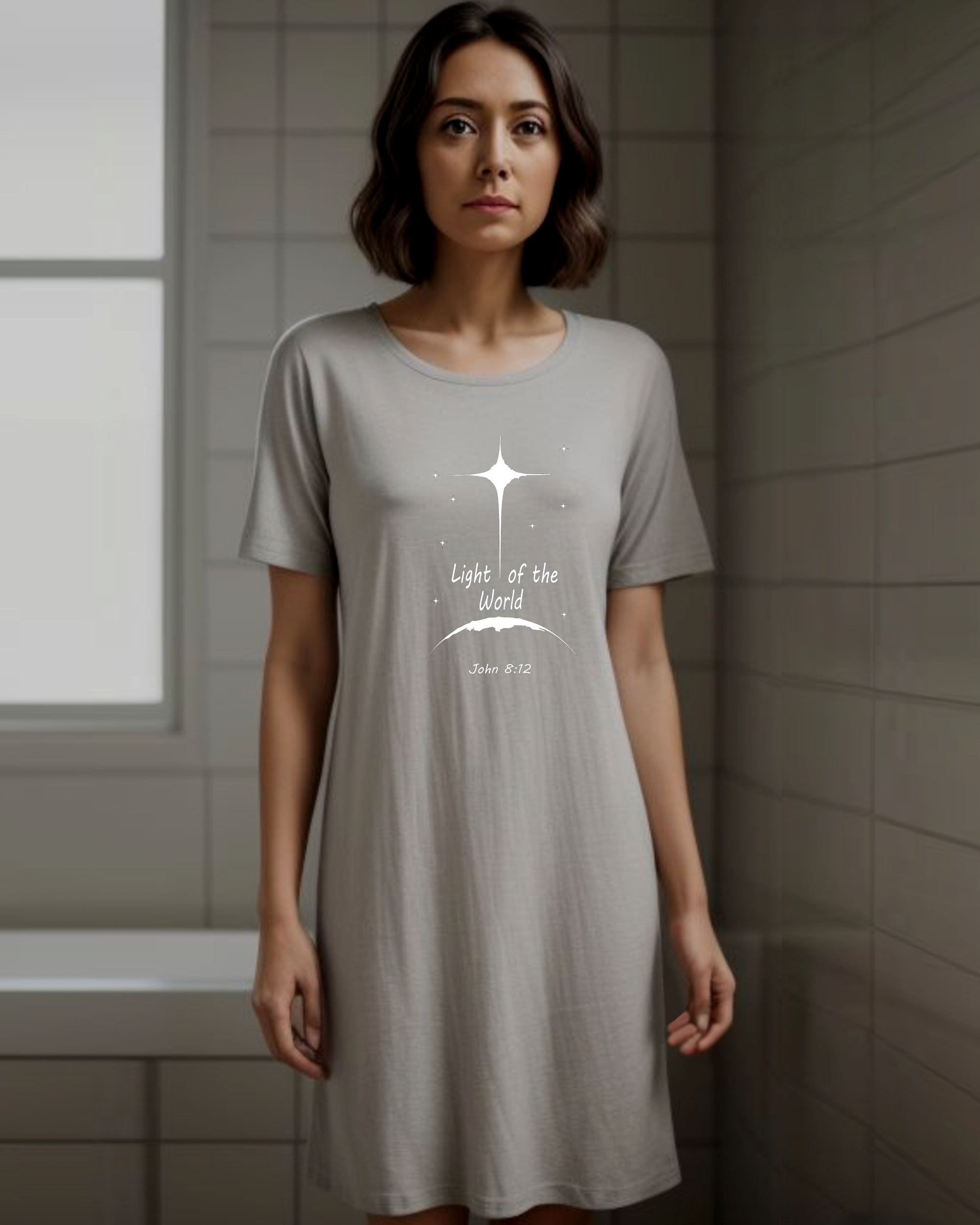 A woman standing in a bathroom wearing a grey t-shirt dress with a white Heavenly Dreamwear logo that says "Light of the World" printed on the front.