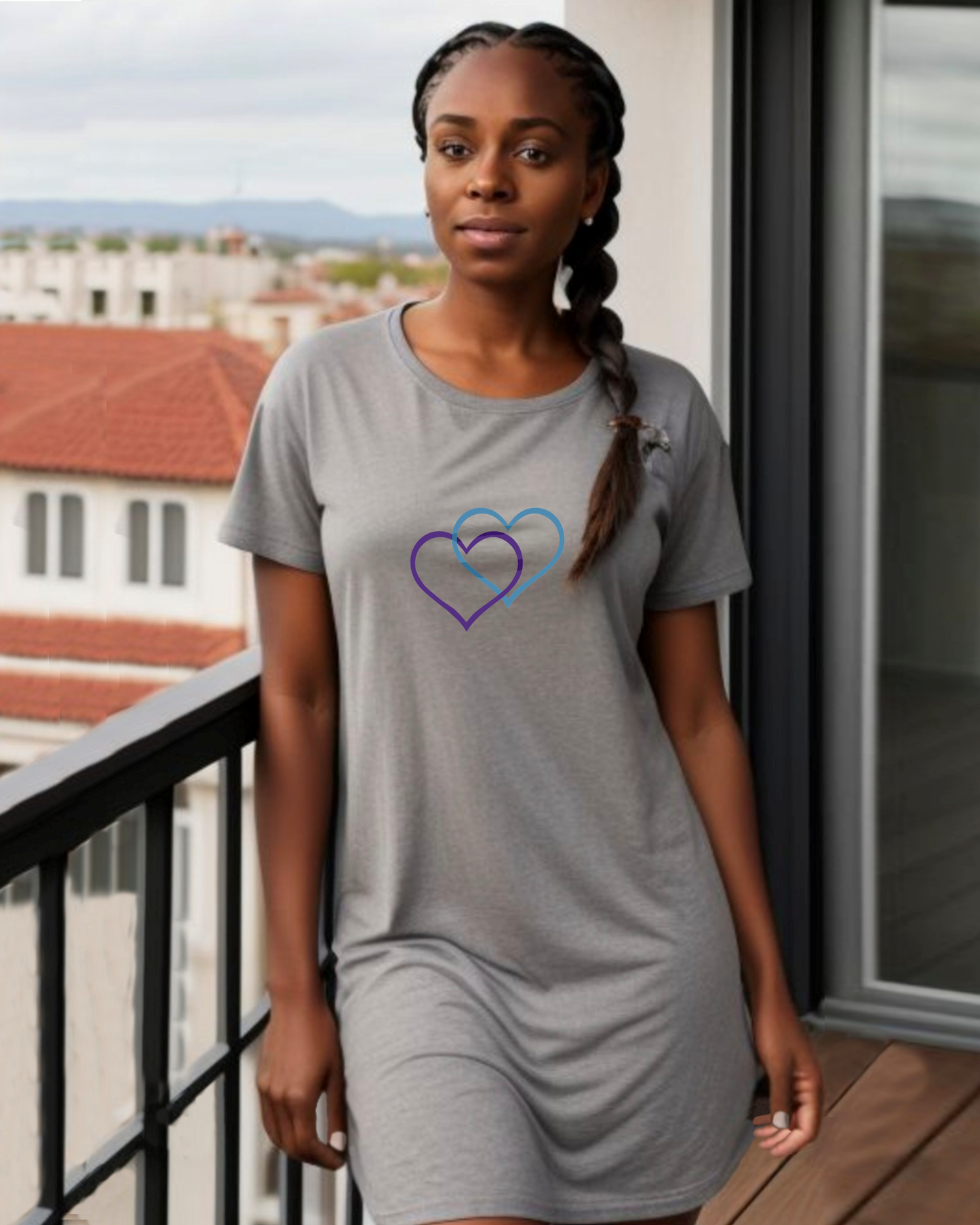 A woman stands on a balcony, dressed in a grey t-shirt dress featuring the Heavenly Dreamwear logo of two intertwined heart shapes printed on the front.