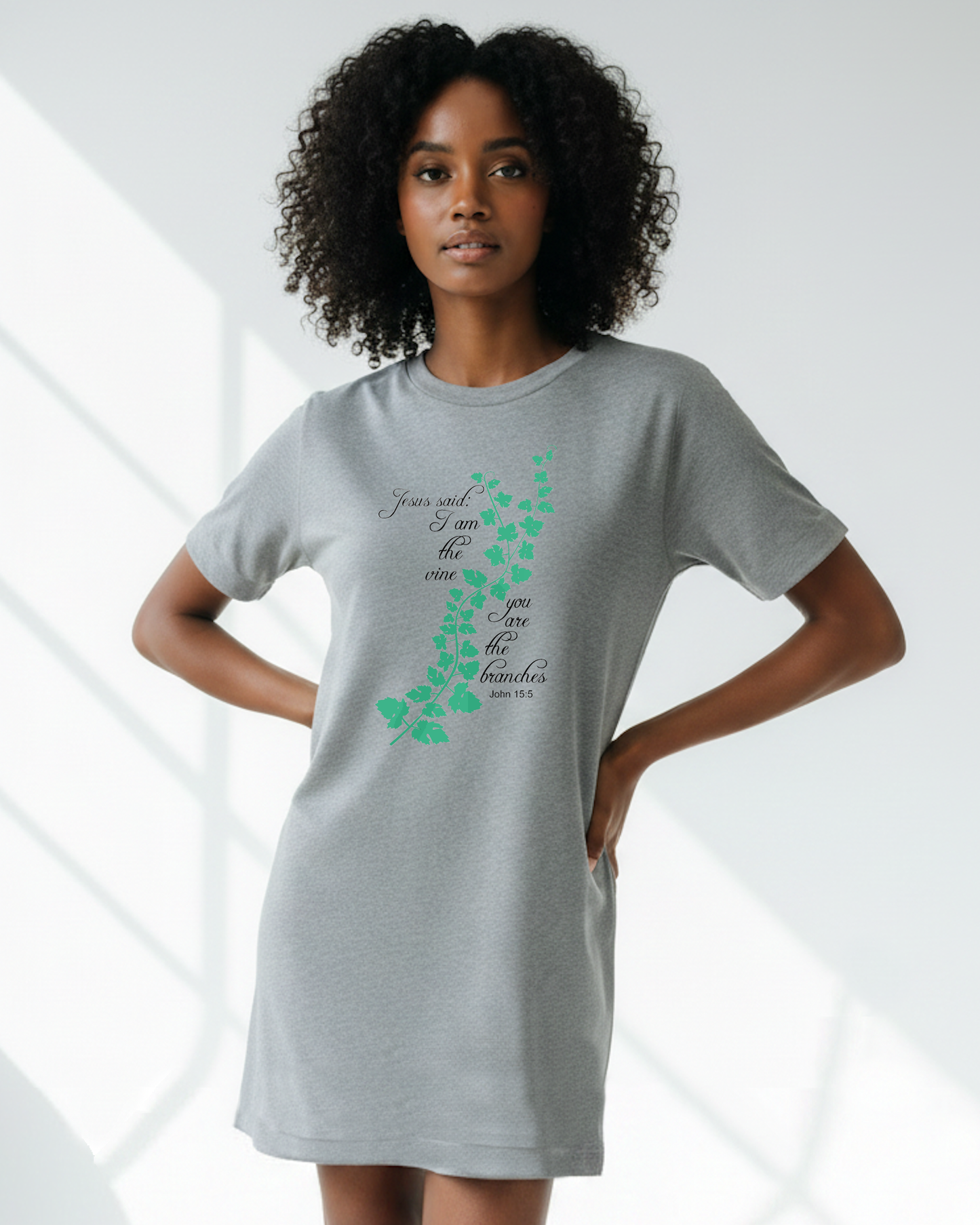 A woman in a bright room wearing a grey t-shirt dress with an elaborate green vine branch with the text, "Jesus said: I am the vine you are the branches" printed in black text on either side.