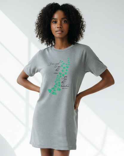 A woman in a bright room wearing a grey t-shirt dress with an elaborate green vine branch with the text, "Jesus said: I am the vine you are the branches" printed in black text on either side.