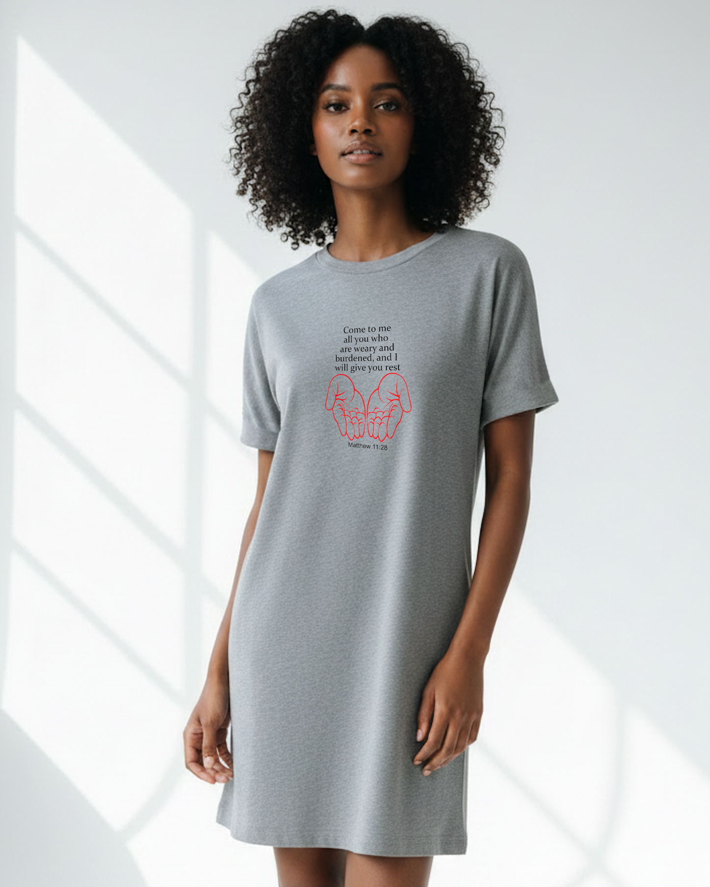 A woman in a bright room wearing a grey Heavenly Dreamwear t-shirt dress with black "come to me all you who are weary" text with two red hands facing palms up logo printed on the front.
