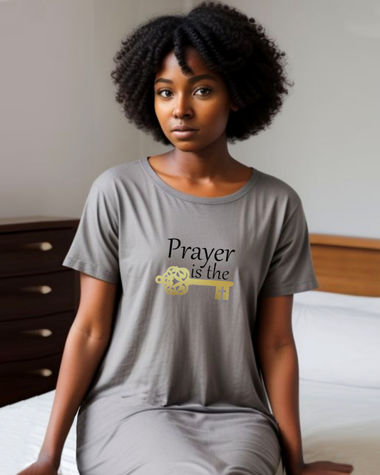 A woman wearing a grey T-shirt dress featuring a design by Heavenly Dreamwear.  The design includes black text that reads, "Prayer is the," followed by a gold key symbol.