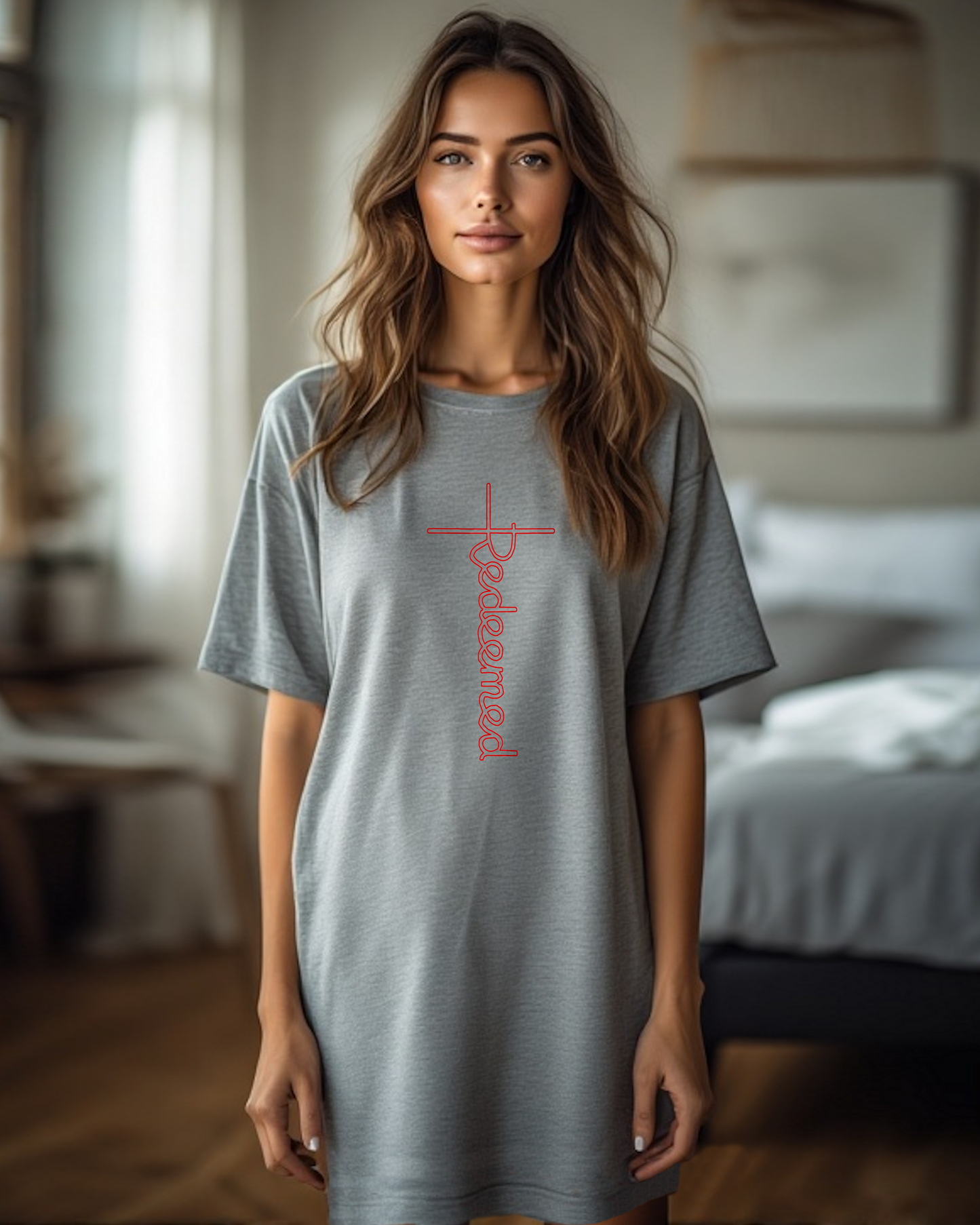 A woman in a bedroom wearing a grey Heavenly Dreamwear t-shirt dress with an elaborate red 'Redeemed' logo printed on the front. 