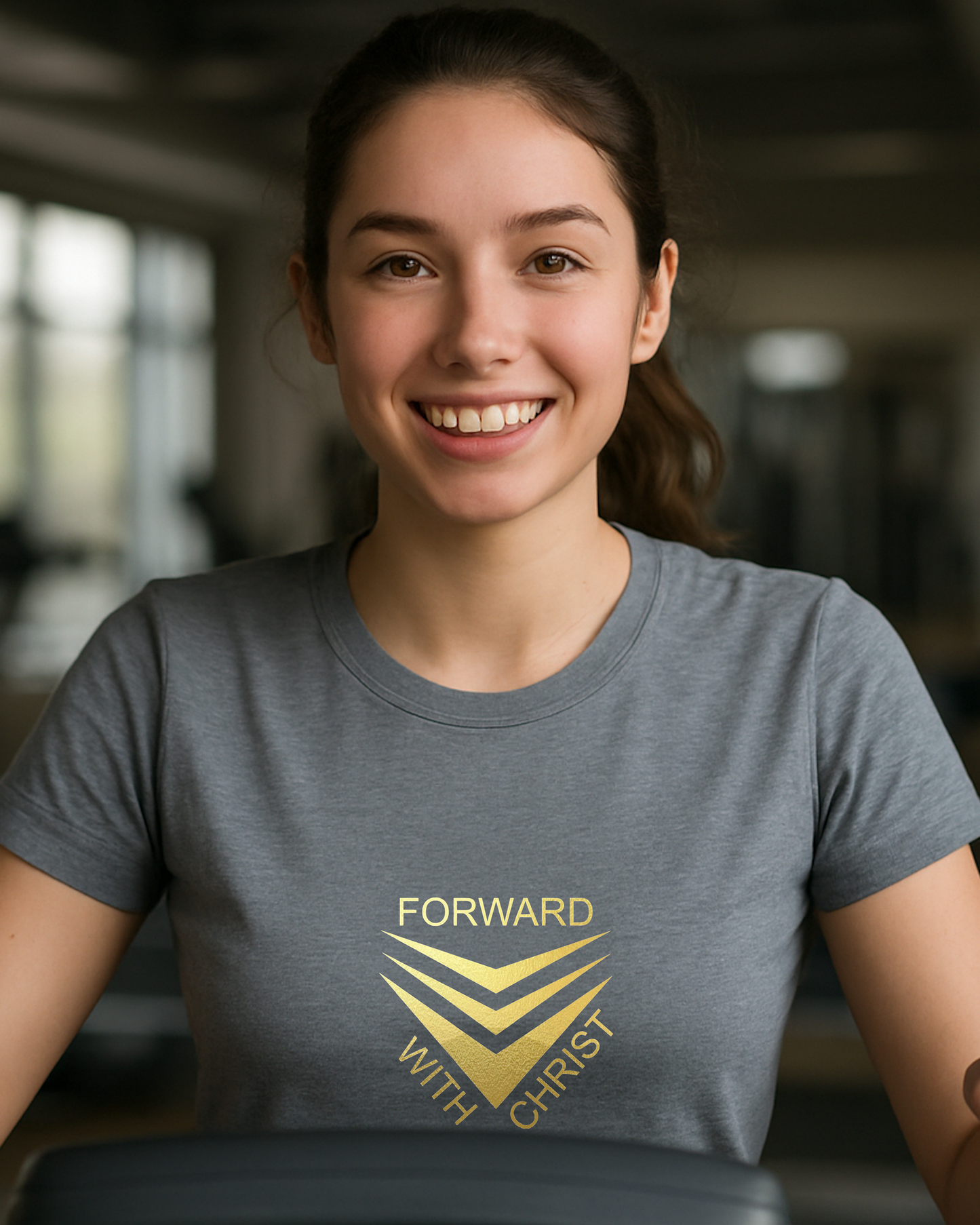 A woman in a gym wearing a grey short sleeve t-shirt that has a gold logo with the words "FORWARD WITH CHRIST" around 3 downward pointing chevrons.