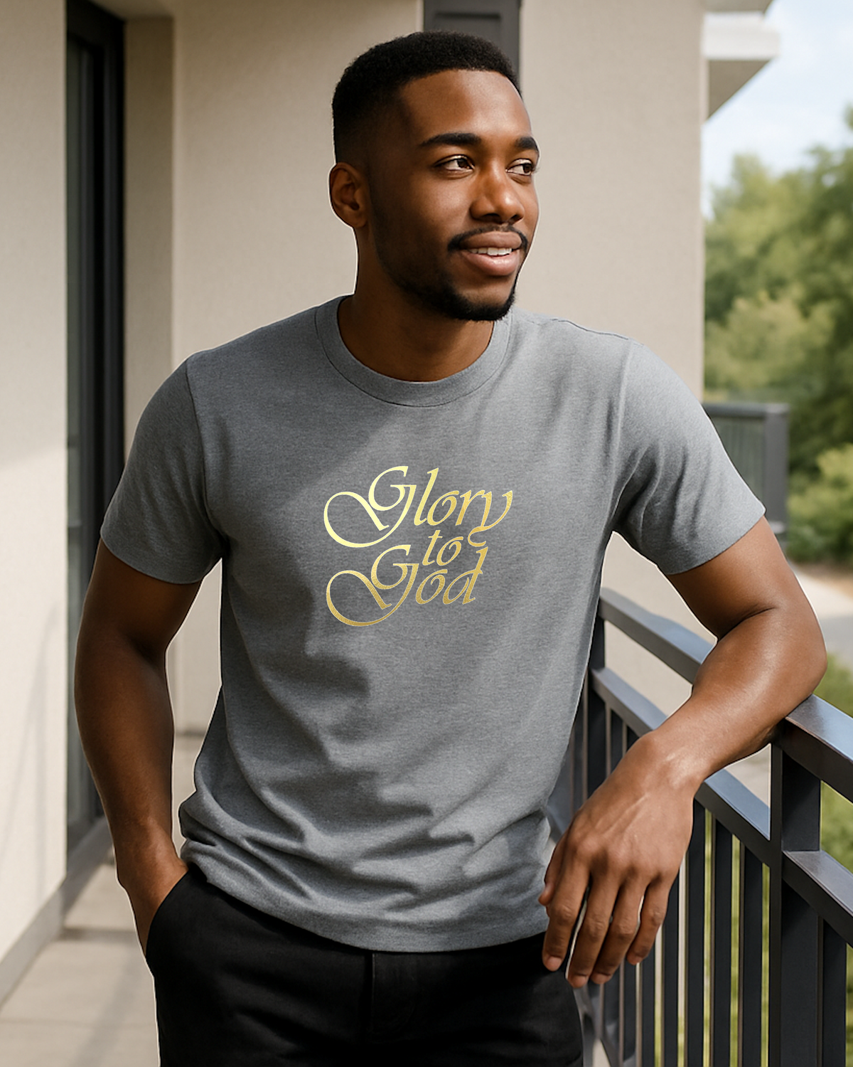 A man wearing a heather grey crewneck t-shirt with 'Glory to God' printed in gold cursive script across the chest, standing on a balcony.