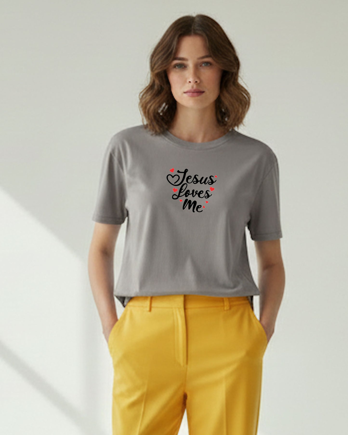 Woman wearing a gray t-shirt with black 'Jesus Loves Me' text with red hearts and yellow pants on a light background
