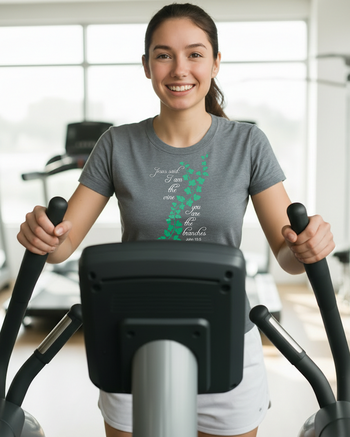 A smiling woman in a gym wearing a grey graphic t-shirt with a green and white vine design and scripture text 'Jesus said I am the vine you are the branches', riding a black and silver elliptical machine.