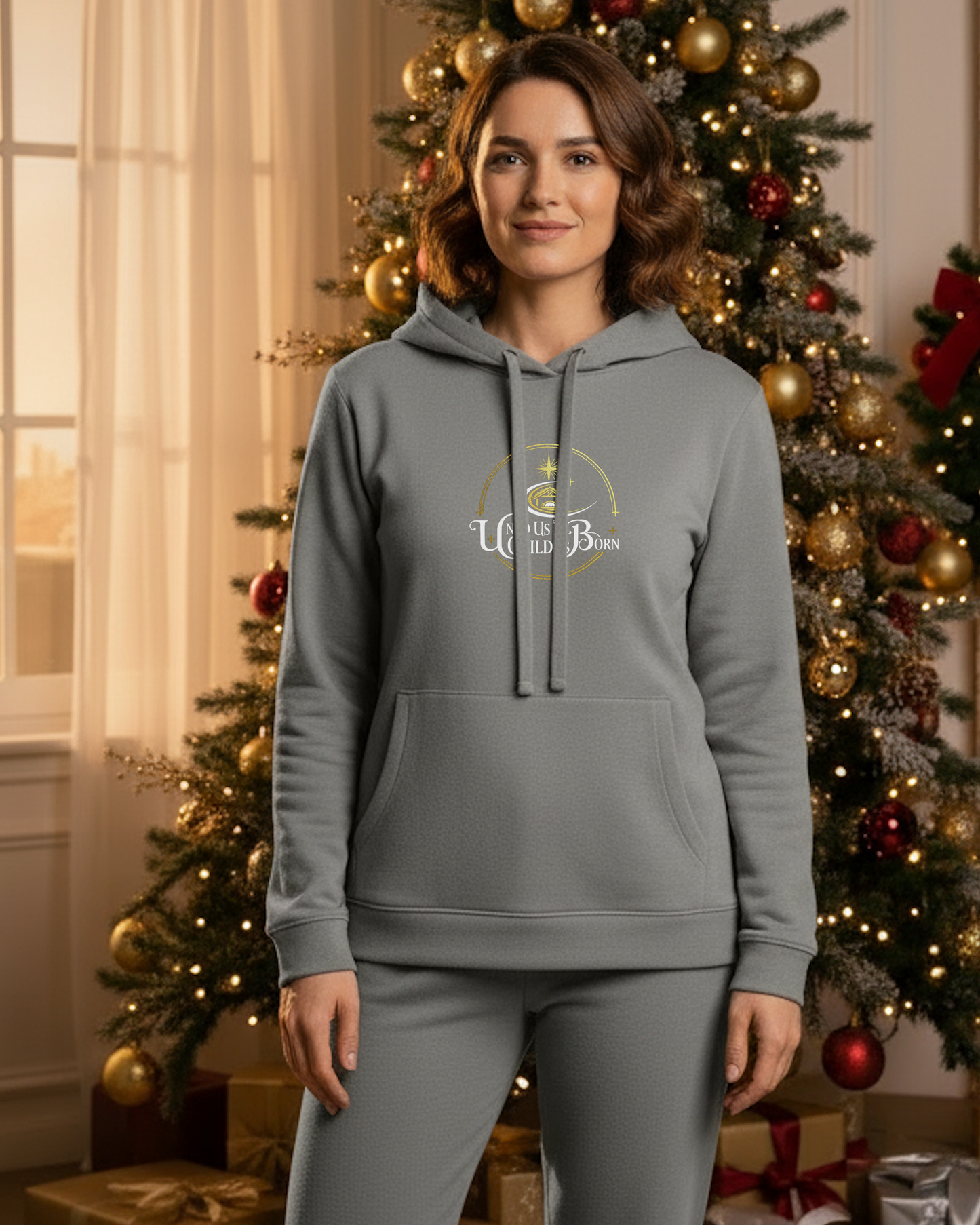 A woman wearing a grey hoodie that has the white text, "Unto Us A Child Is Born" within a gold logo, in Christmas setting room.