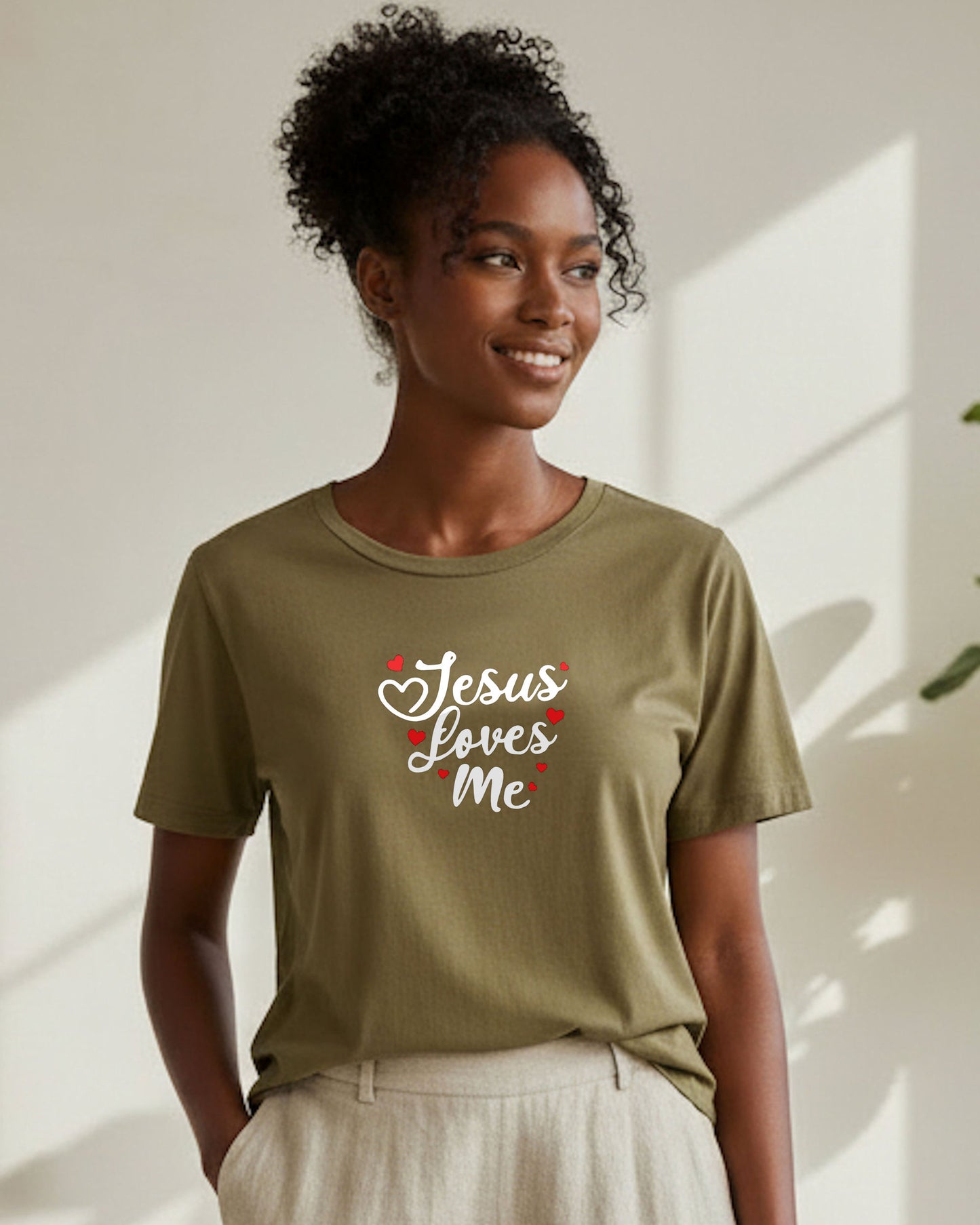 A smiling woman standing in a white room wearing a khaki t-shirt with white text, 'Jesus loves me', with red hearts, and white pants
