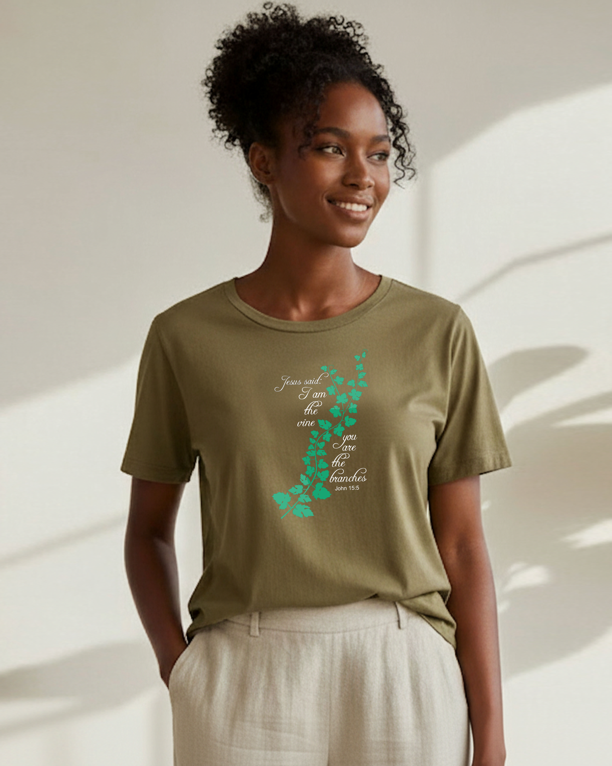 A woman wearing a khaki crewneck t-shirt with a vine graphic logo surrounded by white text 'Jesus said I am the vine you are the branches', paired with light beige linen trousers, set against a warm beige wall with soft leaf shadows.