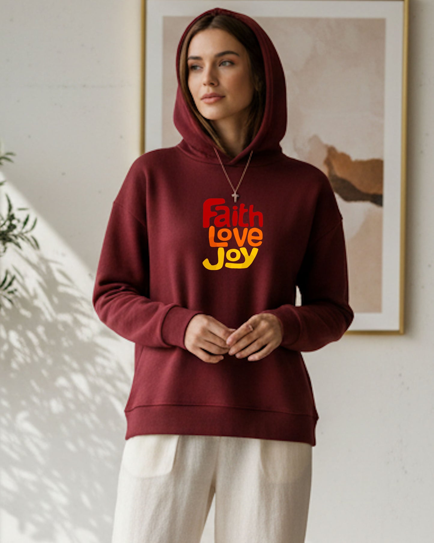 Woman standing indoors wearing a maroon hoodie with a red, orange and yellow Heavenly Dreamwear text logo printed on the front.