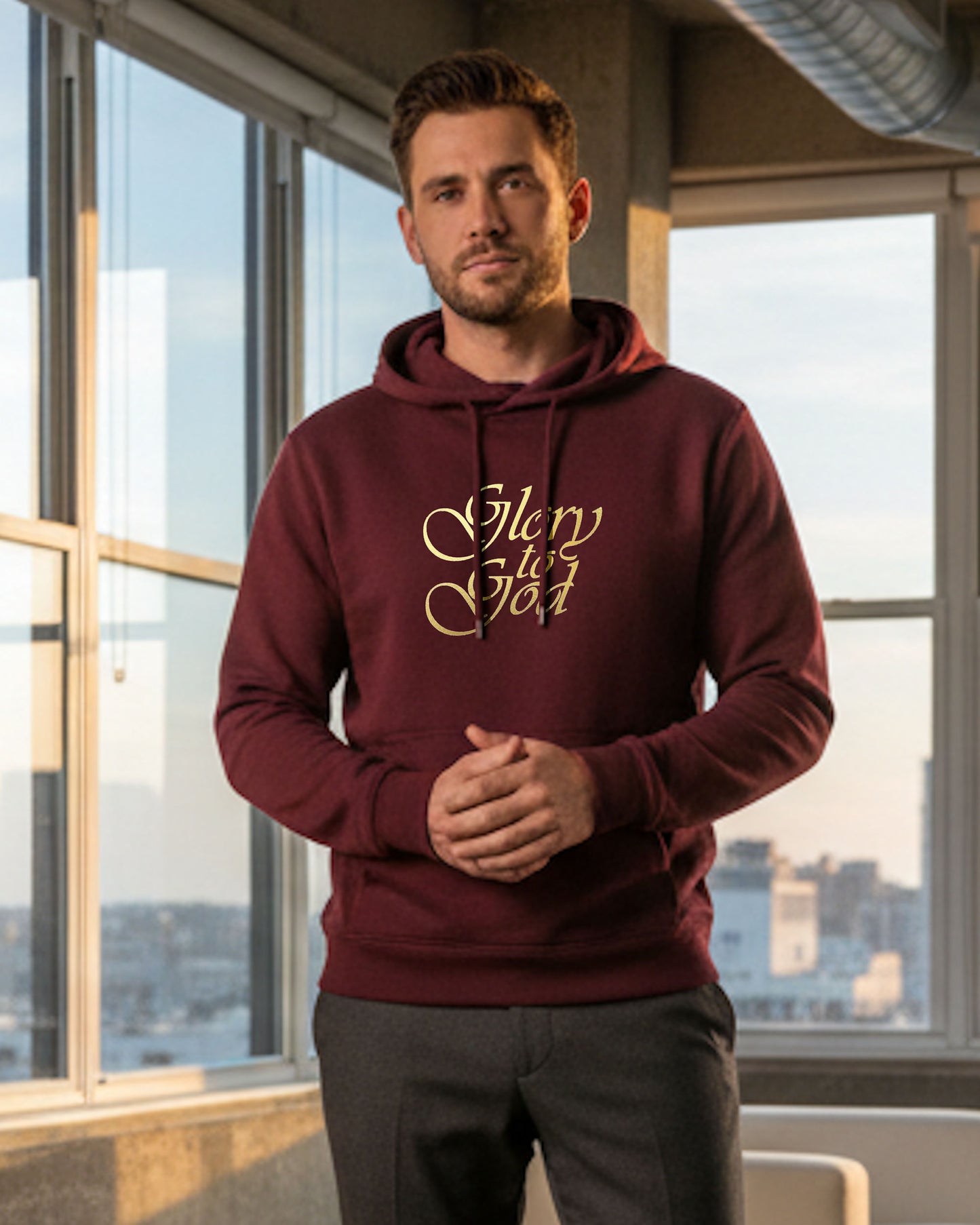 Man standing indoors wearing a maroon hoodie with a gold Heavenly Dreamwear "Glory to God" text logo printed on front. 