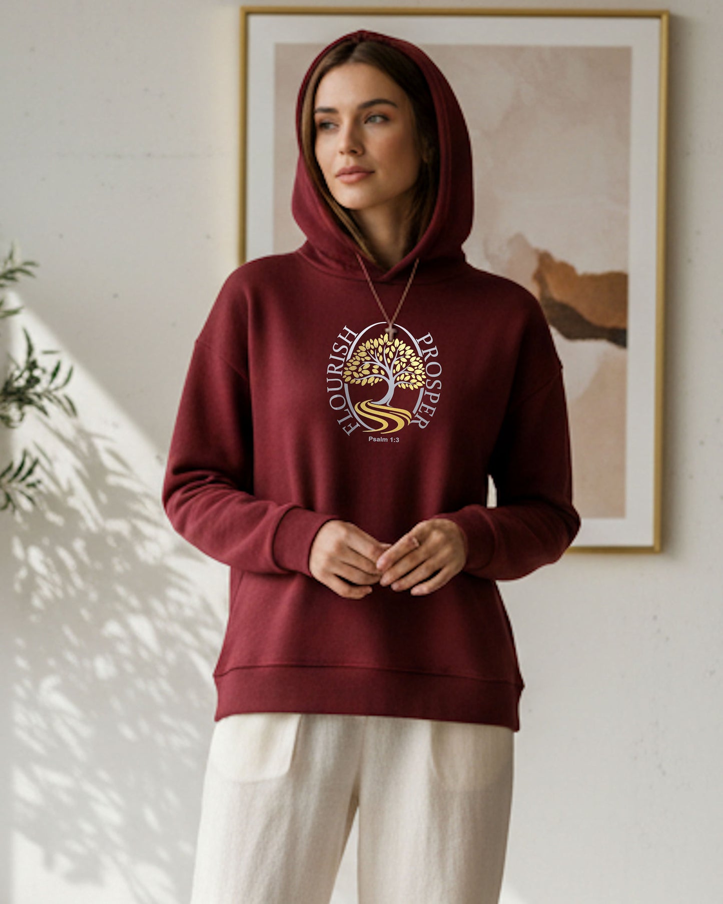 Woman indoors wearing a maroon hoodie with a gold/silver logo with text 'Flourish Prosper Psalm 1:3'