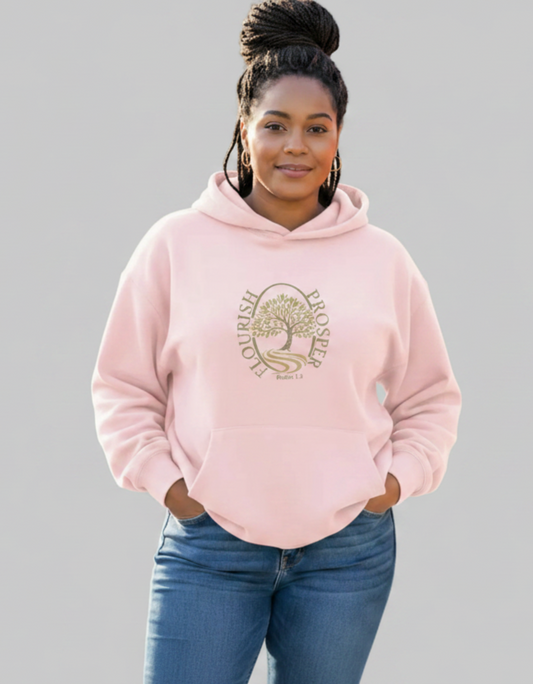 A woman wearing a light pink pullover hoodie with a gold-toned 'FLOURISH PROSPER' tree graphic on the chest, paired with blue jeans, standing against a pale grey background.