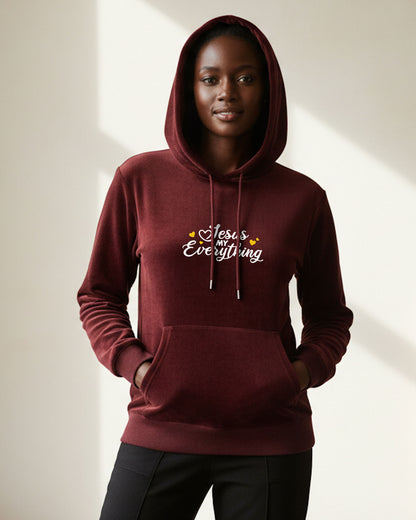 Woman wearing a maroon hoodie with 'Jesus loves everything' white text with yellow hearts on a neutral background