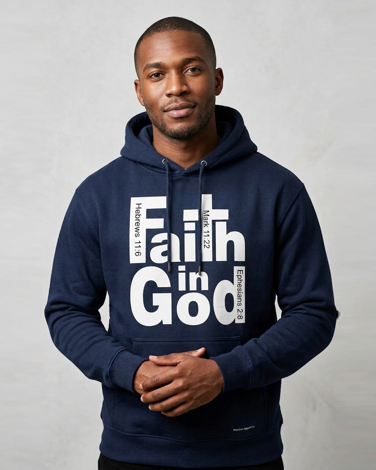 A man wearing a navy blue hoodie with a large white graphic print that reads 'Faith in God' with scripture references, standing against a light gray textured background.