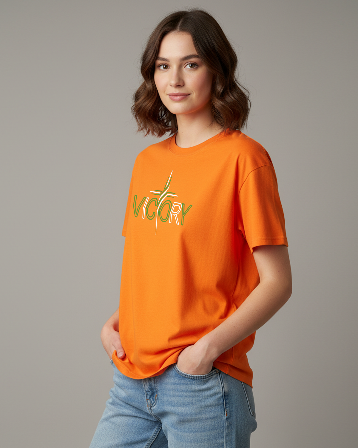 A woman wearing a bright orange short-sleeve t-shirt with a green and white 'VICTORY' graphic on the chest, paired with light blue jeans, photographed against a solid gray studio background.