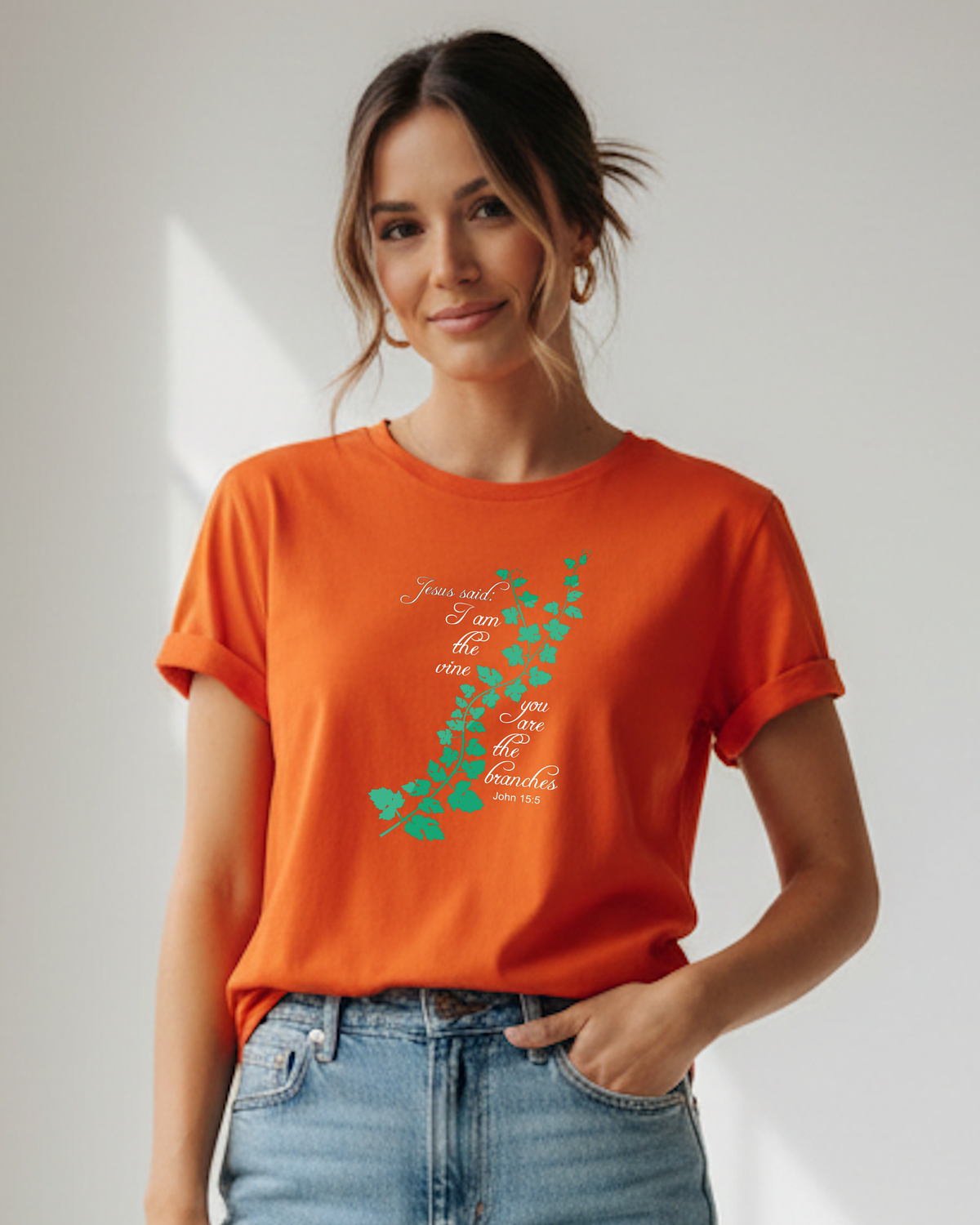 A woman wearing an orange short-sleeve crewneck t-shirt with a green vine graphic and white cursive text 'Jesus said I am the vine you are the branches', paired with light blue jeans, standing in a brightly lit room with soft shadows.