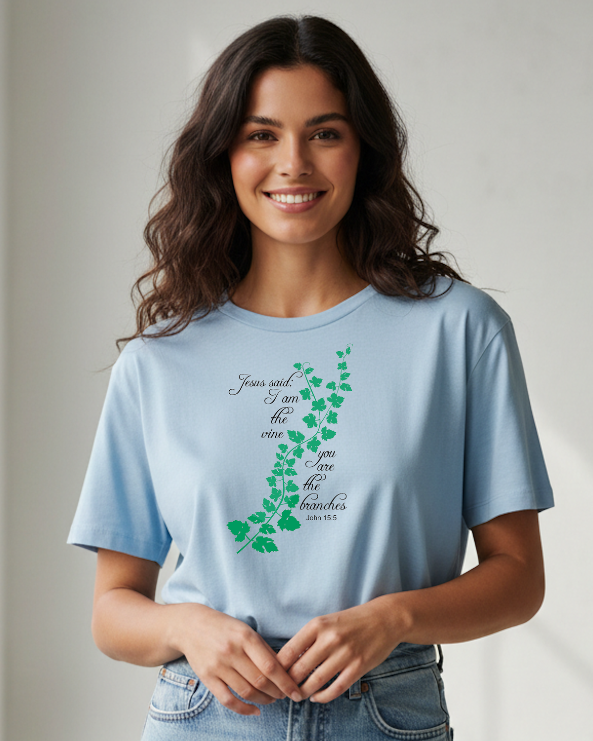 A woman wearing a light blue graphic t-shirt with a green ivy vine and cursive text reading 'Jesus said I am the vine, you are the branches' (John 15:4-5), paired with blue jeans, set against a neutral light grey background.